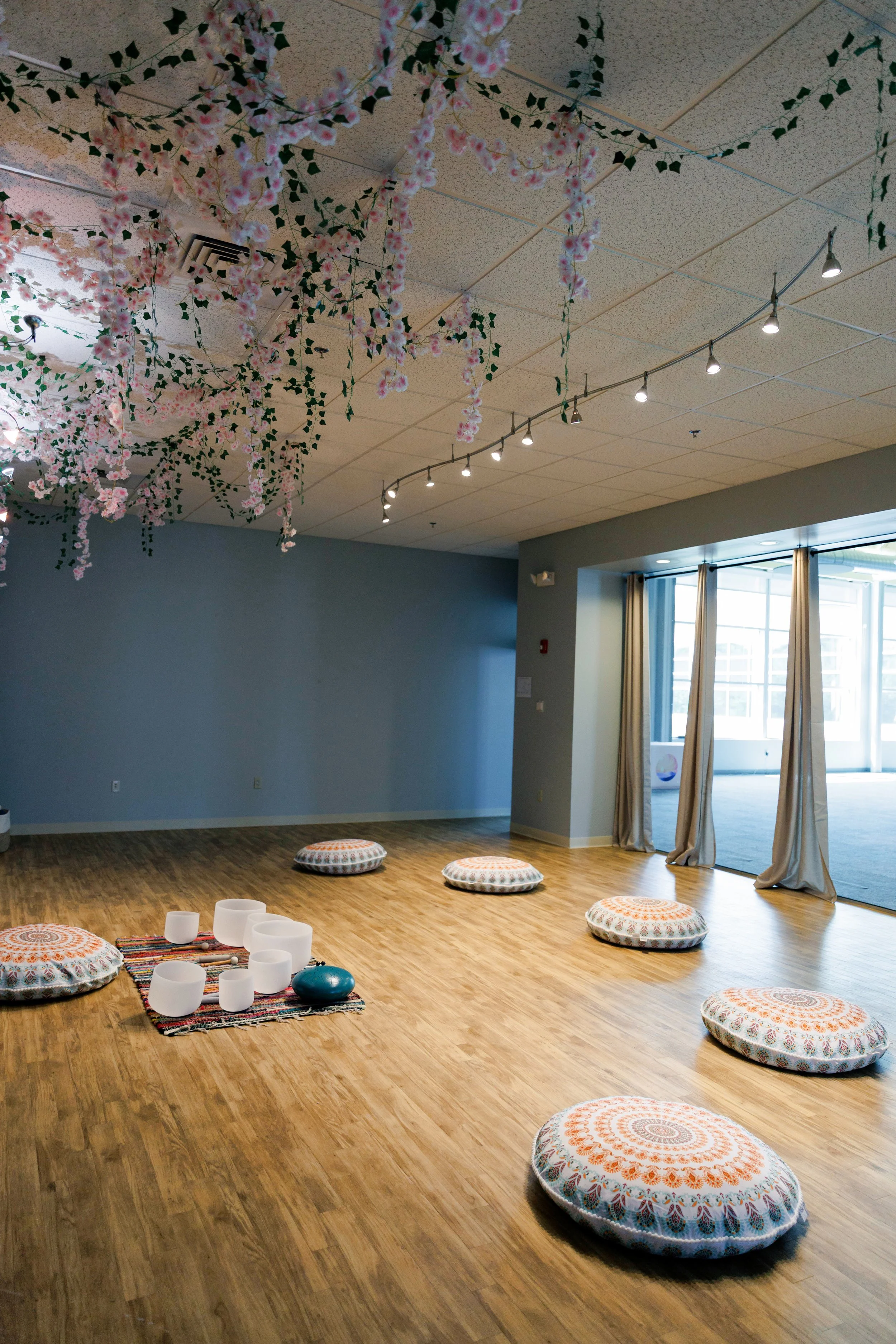 A room decorated with pink cherry blossom hanging from the ceiling with green ivy garlands. The floor has round patterned cushions and some white singing bowls on a colorful woven mat near the wall with windows. The room appears suitable for meditation or yoga sessions.