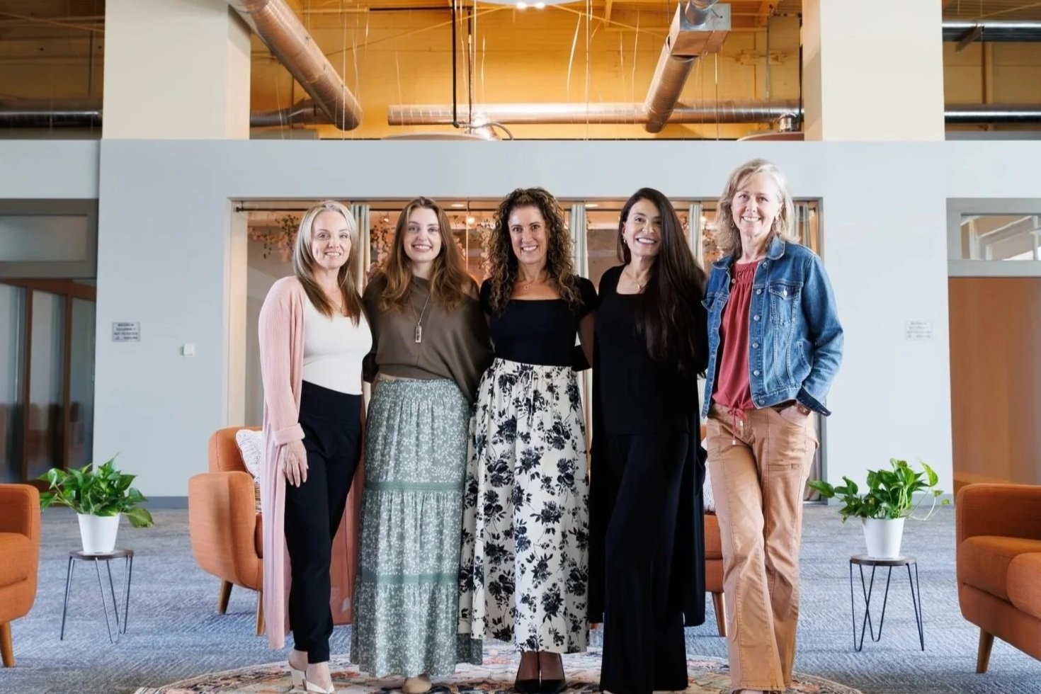 Five women standing together in an indoor office or lounge area, smiling for the camera. The background features modern decor, green potted plants, and orange chairs.