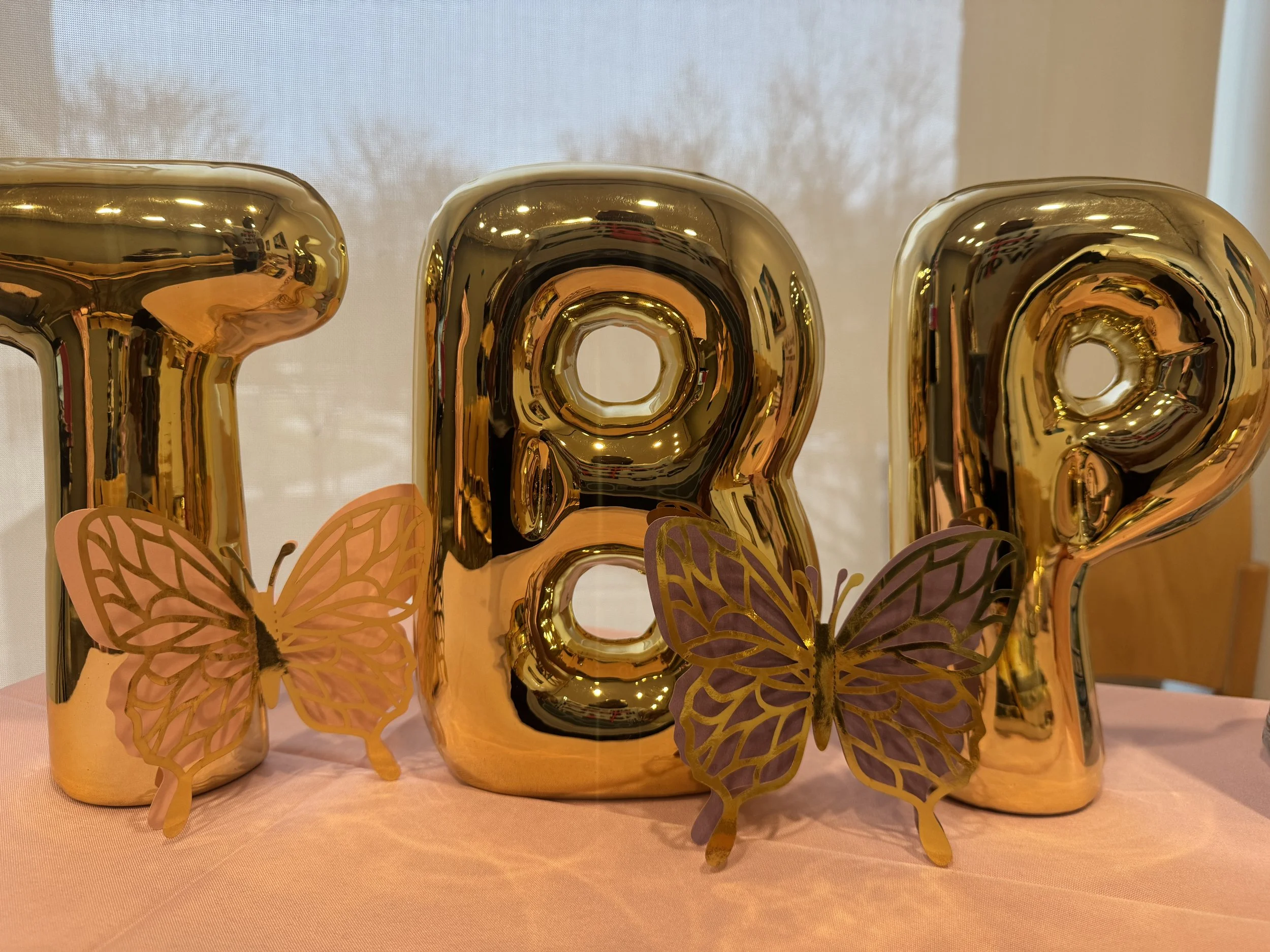 Gold balloons spell out 'TAP' with pink and purple butterfly decorations in front, on a pink tablecloth.