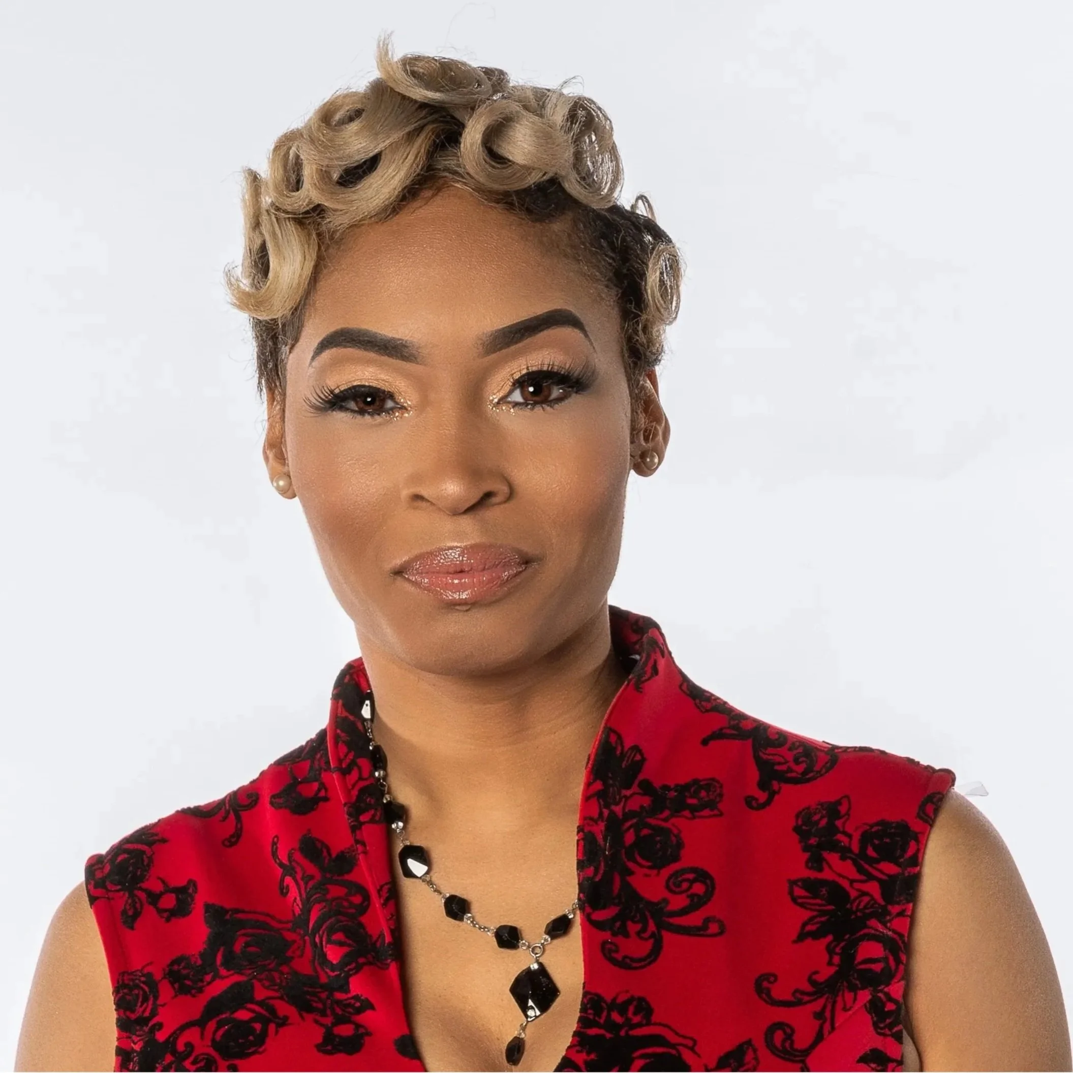Portrait of a woman with styled blonde curls, wearing a red sleeveless top with black floral patterns, and a black beaded necklace with a large black pendant, standing against a plain white background.