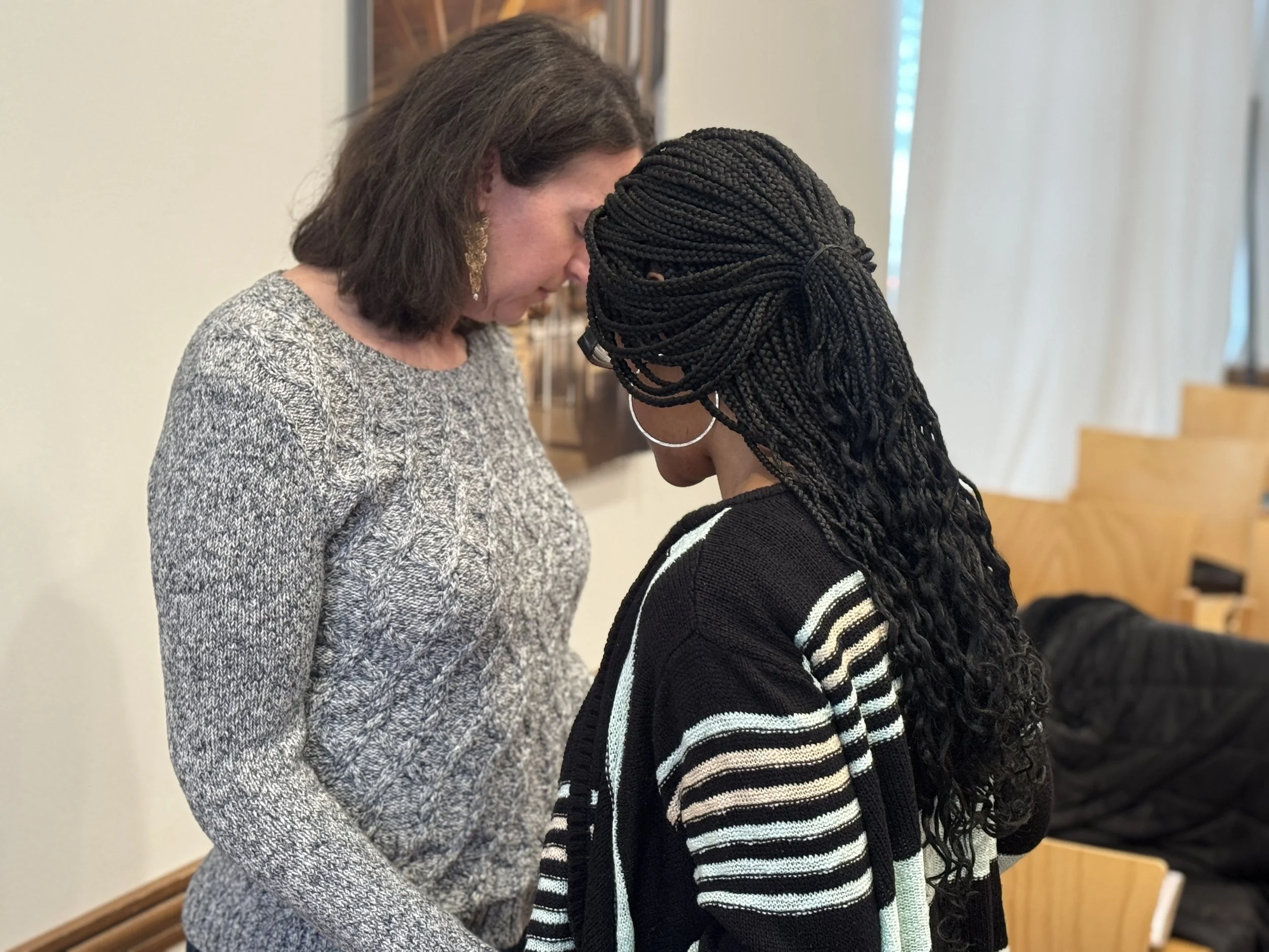 Two women standing close with foreheads touching, engaged in a heartfelt moment, indoors.