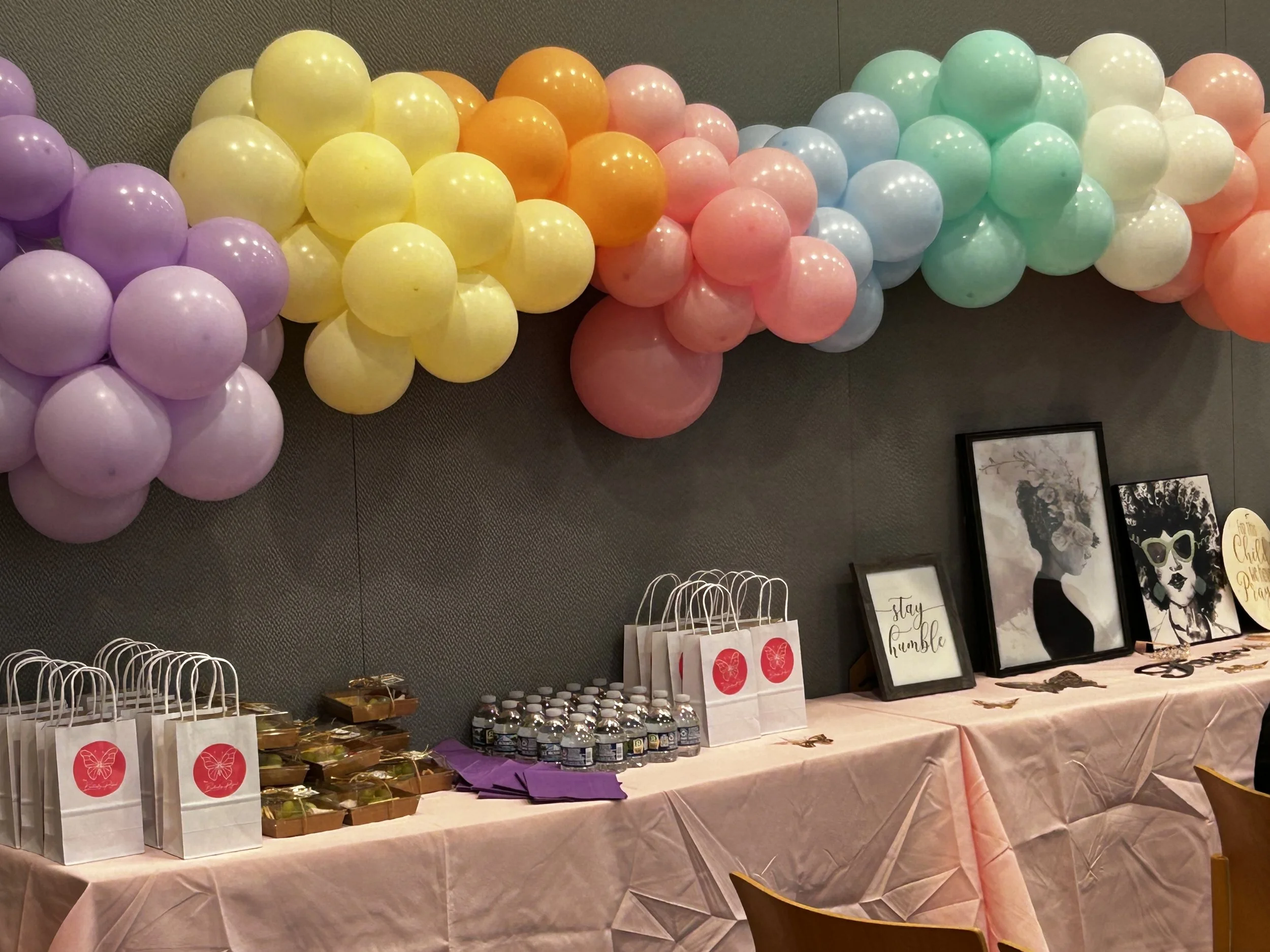 Colorful pastel balloon garland with purple, yellow, orange, pink, blue, green, and white balloons decorates the wall above a table. The table is covered with a pink tablecloth and holds gift bags, water bottles, framed artwork, and decorative signs.