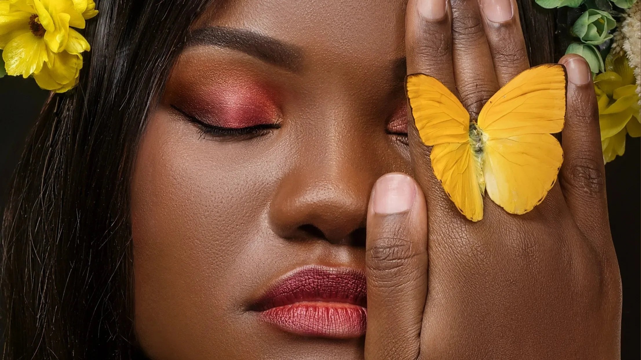 A woman with dark skin and closed eyes, makeup with pink and red eyeshadow, pink lipstick, is touching her face with her left hand. A yellow butterfly rests on her hand, and yellow flowers are visible in her hair.
