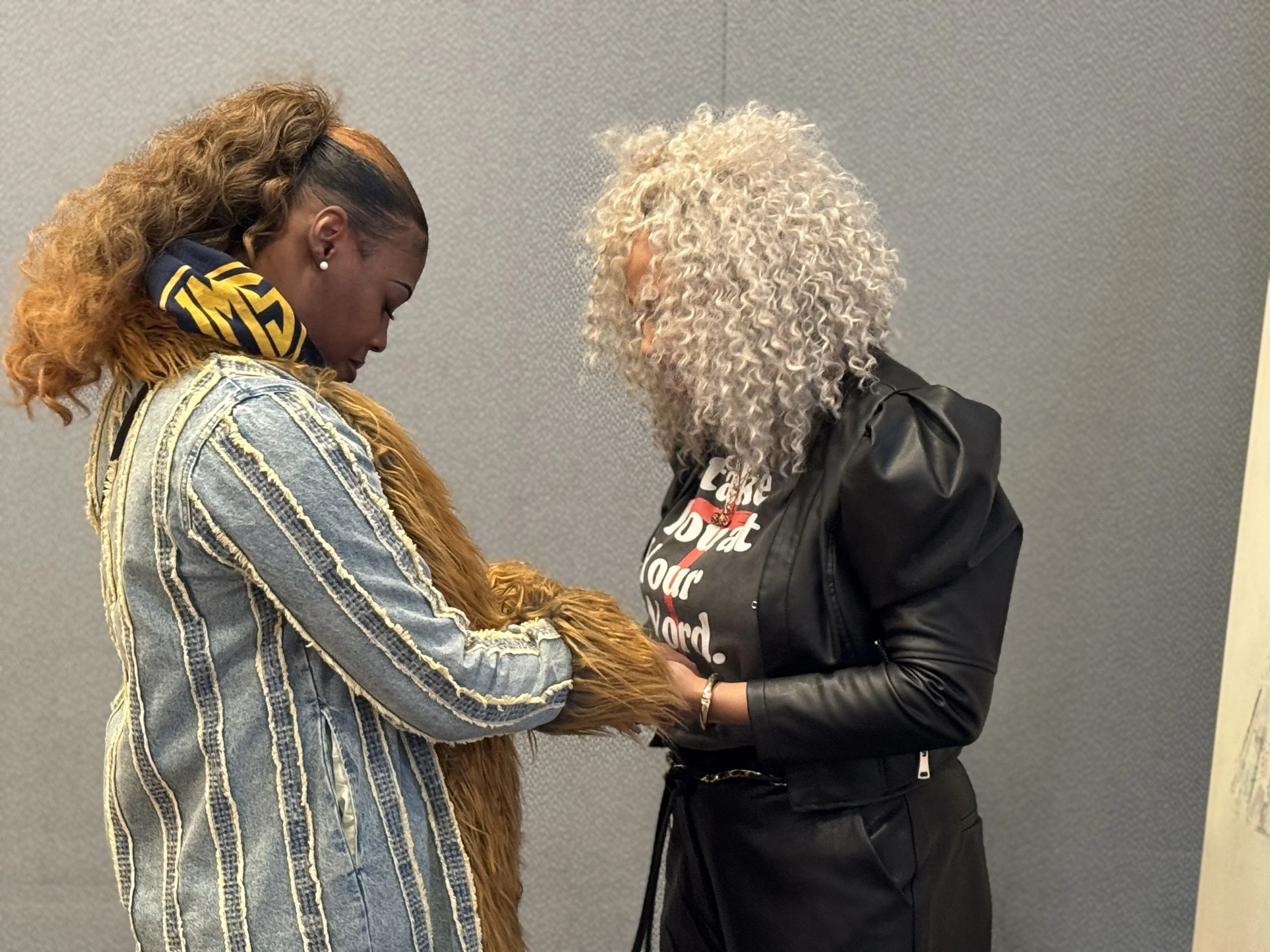 Two women standing close together, one with long curly gray hair and the other with long brown hair, are looking down at something they are holding in their hands.