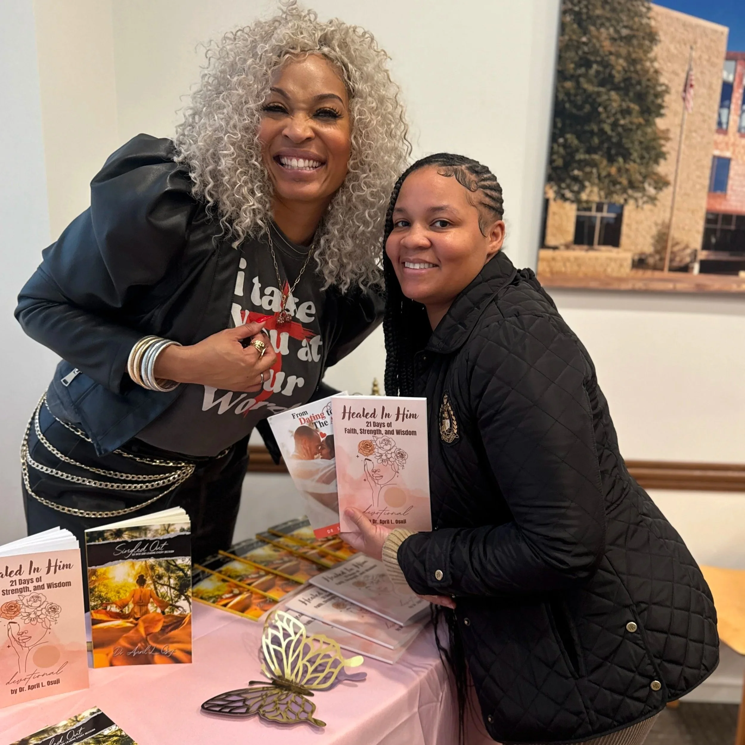 Two women smiling at a book signing event, holding copies of a book titled 'Healed In Him: 21 Days of Faith, Strength, and Wisdom' by Dr. April L. Osuji, with books displayed on a table in front of them.