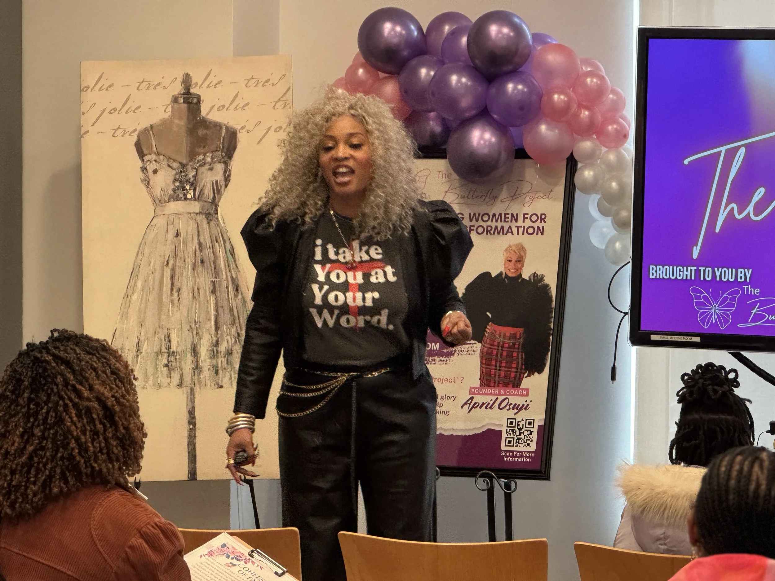 A woman with curly blond hair is speaking at a conference or event, wearing a black blazer and a McDonald's-themed t-shirt that says "take You at Your Word." She has multiple bracelets on her wrist. In the background, there is a dress poster and a butterfly-themed banner, with balloons in shades of purple, pink, and white forming an arch above her. There are audience members seated and listening.