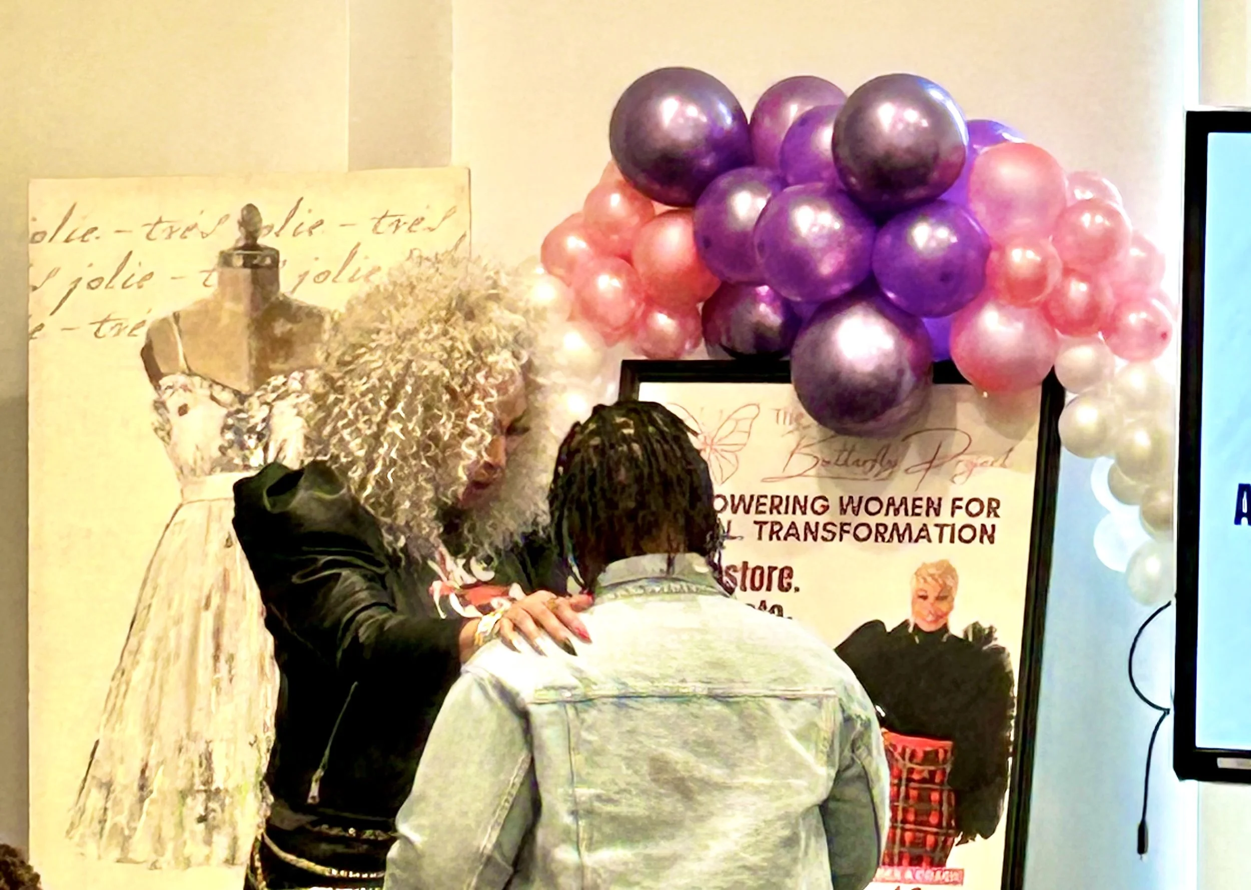 Two women engaged in a conversation at an event, standing in front of a backdrop decorated with pink, purple, and white balloons and a banner with text that mentions empowering women for social transformation.