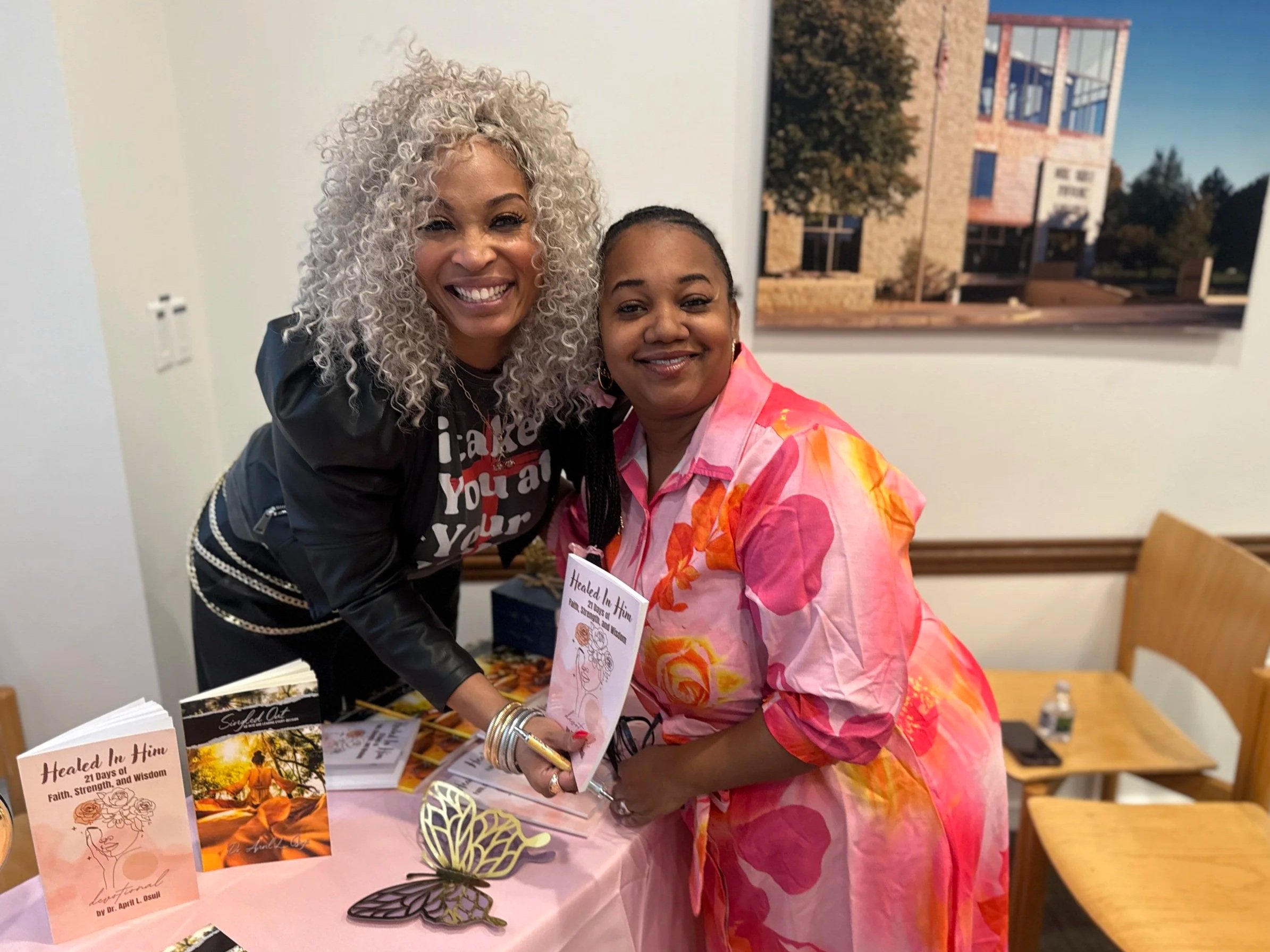 Two women smiling at a book signing event, with books on the table titled 'Healed In Him.' One woman has curly gray hair and is wearing a black jacket, while the other has dark hair pulled back and is wearing a pink and orange floral blouse. They are holding a copy of the book together.