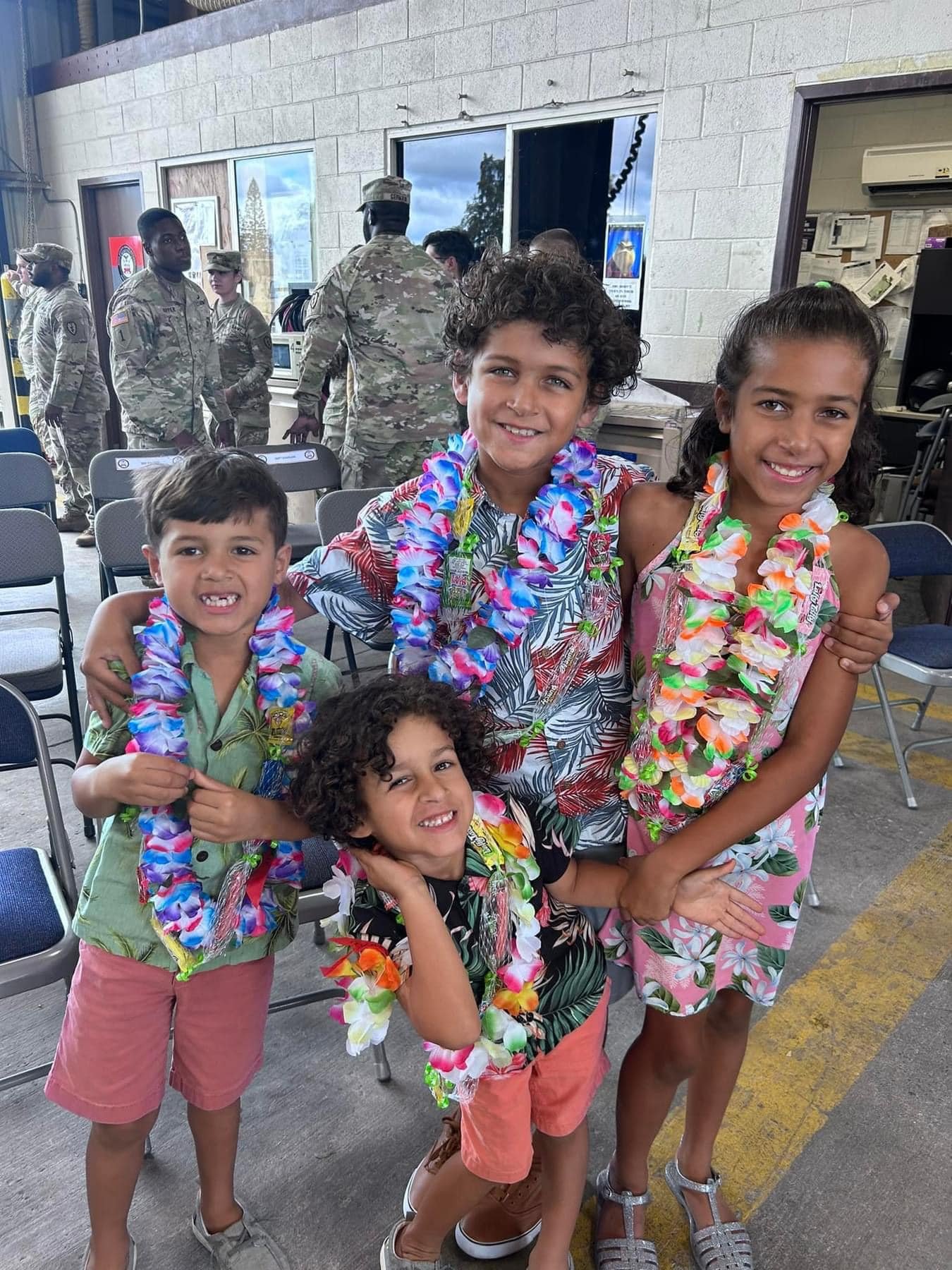 Group of children and a woman smiling, wearing colorful leis, in a space with chairs and military personnel in the background.