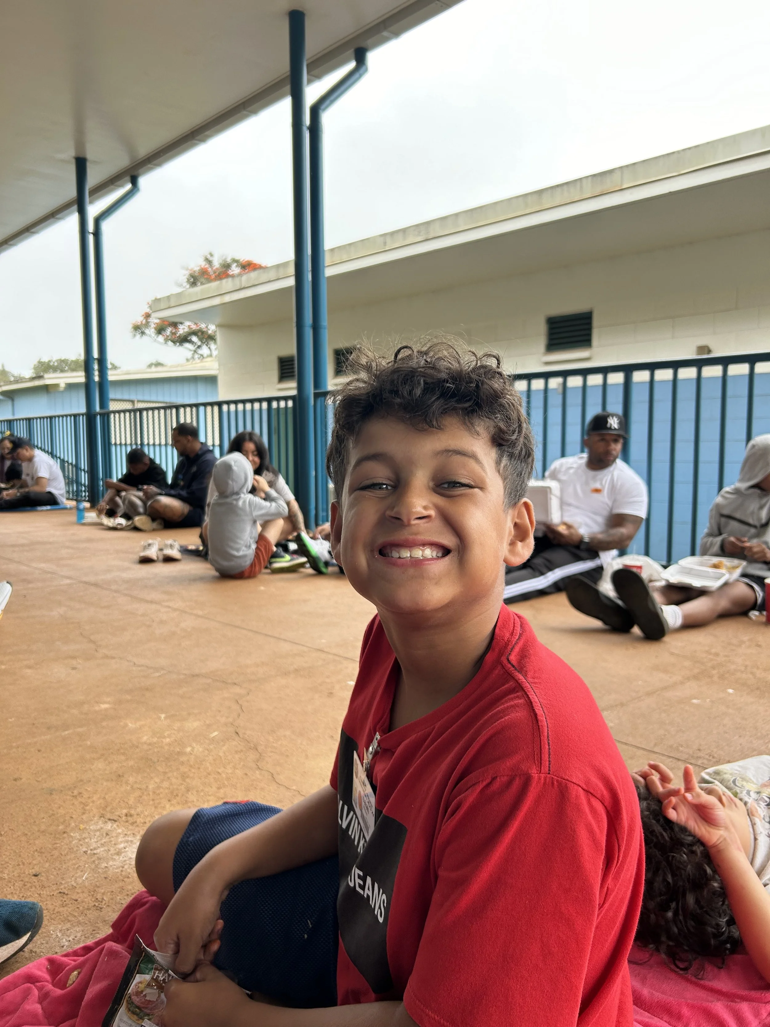 A young boy with curly hair smiling at the camera, sitting on a blanket in an outdoor covered area with other people sitting and eating in the background.