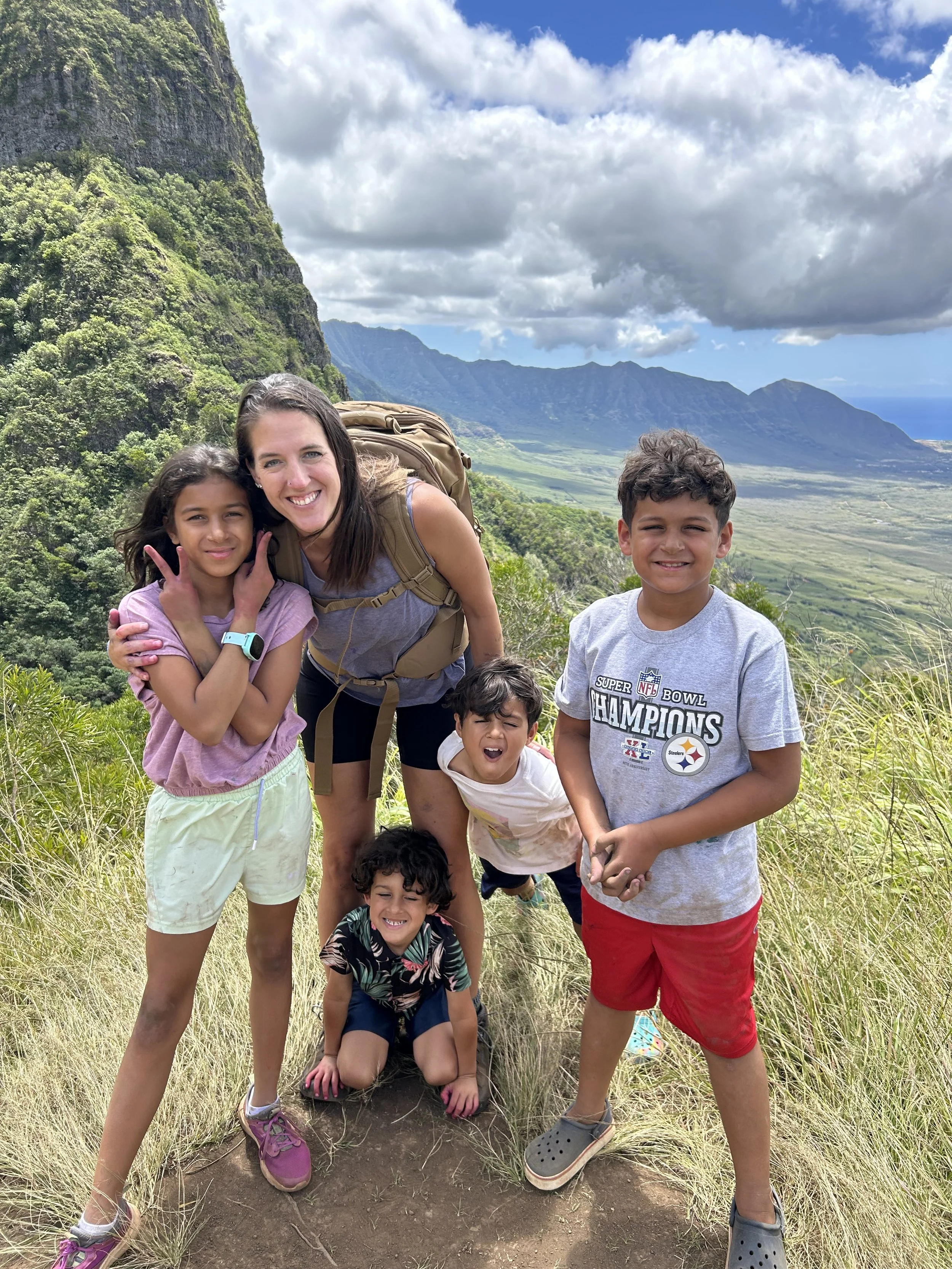Group of six children and one adult on a mountain trail with lush green hills, mountains, and partly cloudy sky in the background.