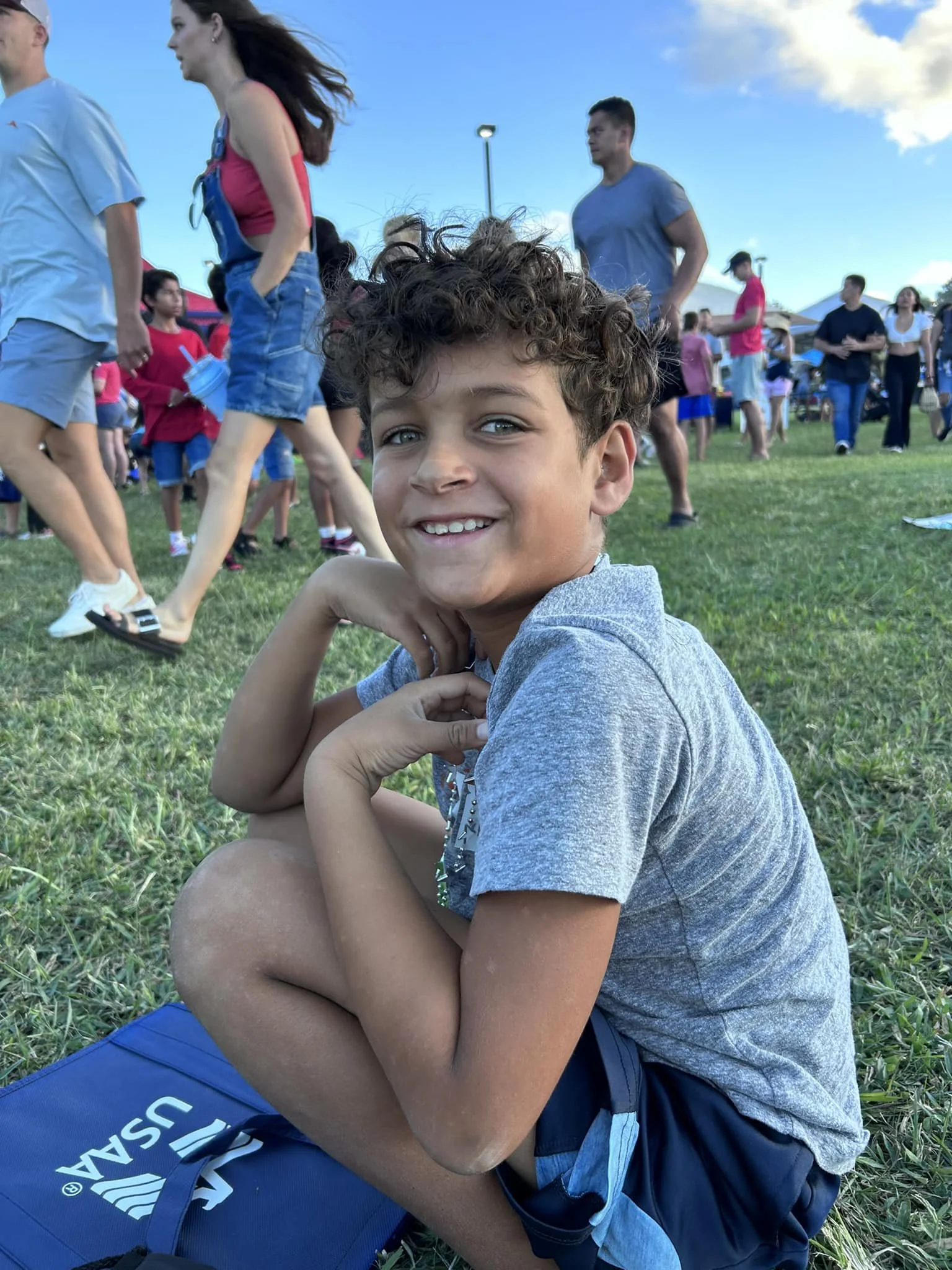 A smiling boy with curly hair sitting on grass during an outdoor event with many people in the background.