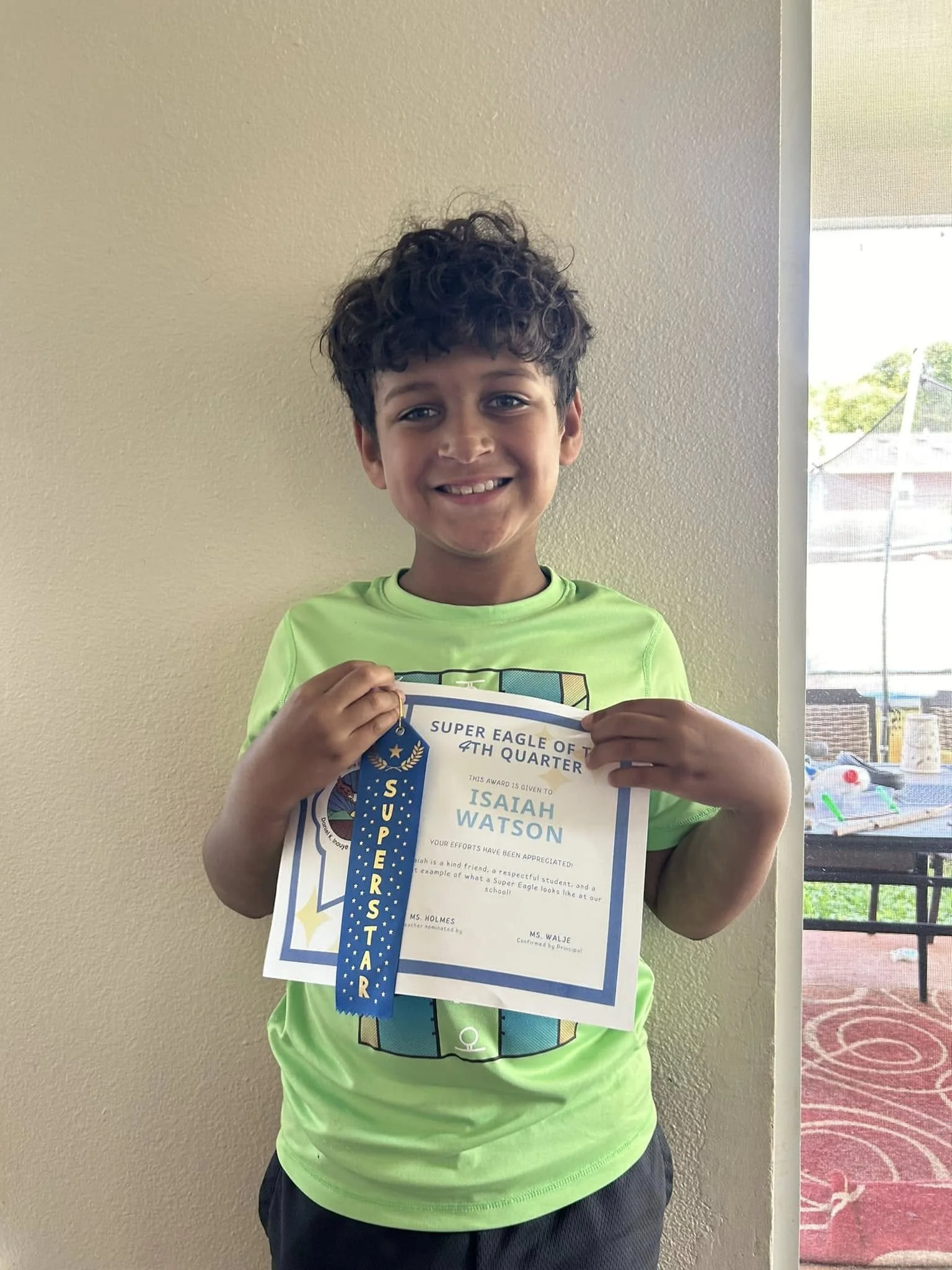 A young boy with curly dark hair smiling and holding a certificate and a blue ribbon that says 'Superstar' in front of a beige wall and a door open to an outdoor area with a table and a red carpet.