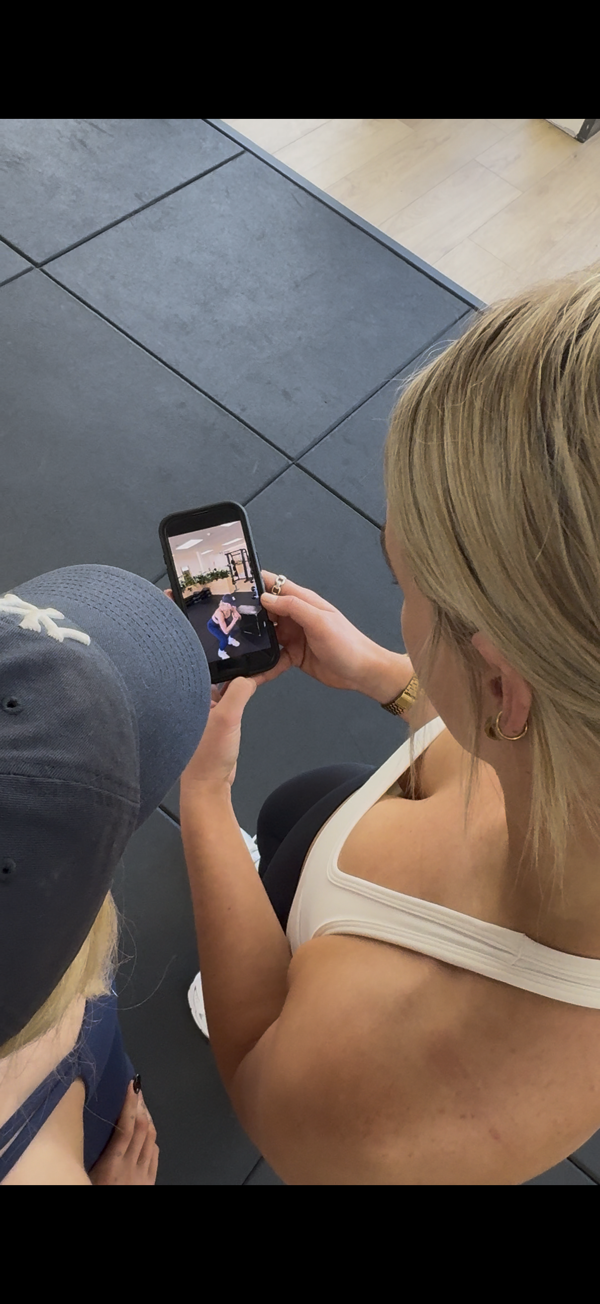 Woman in a white tank top taking a photo with her phone, kneeling on the floor with a young girl in a baseball cap nearby, on a black tiled floor in a gym or fitness center.
