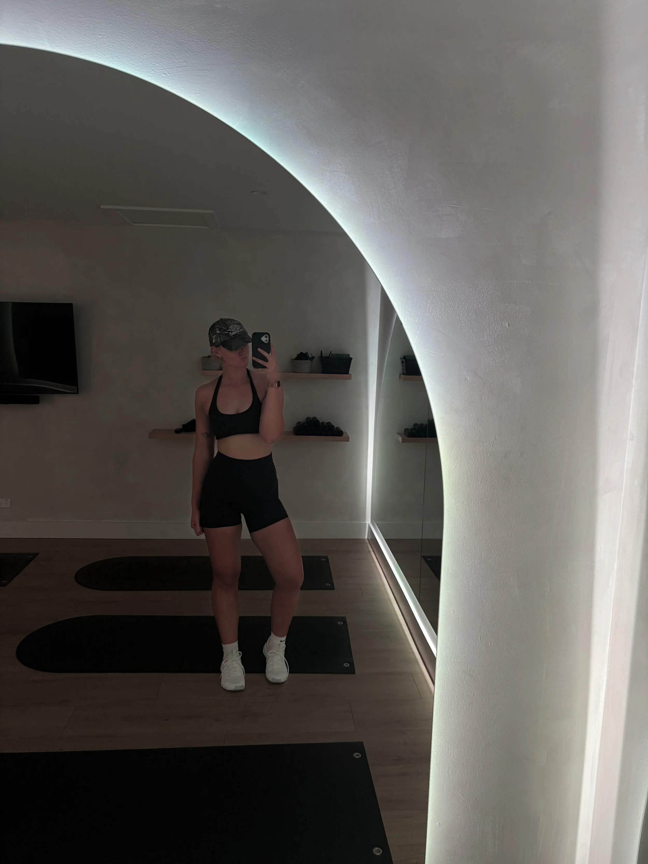 A woman taking a mirror selfie in a room with wooden floors, black exercise mats, and wall-mounted shelves with exercise equipment. She is wearing a sports bra, shorts, sneakers, and a patterned cap, and standing in front of a curved mirror.