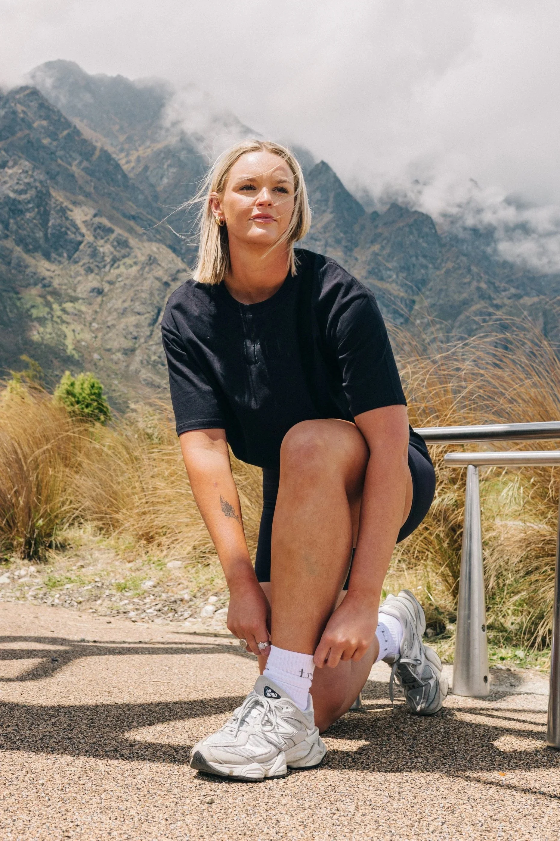 A young woman with blonde hair, wearing a black t-shirt, white sneakers, and white socks, is kneeling on one knee near a metal railing in an outdoor mountainous area with tall grass and mountains in the background.