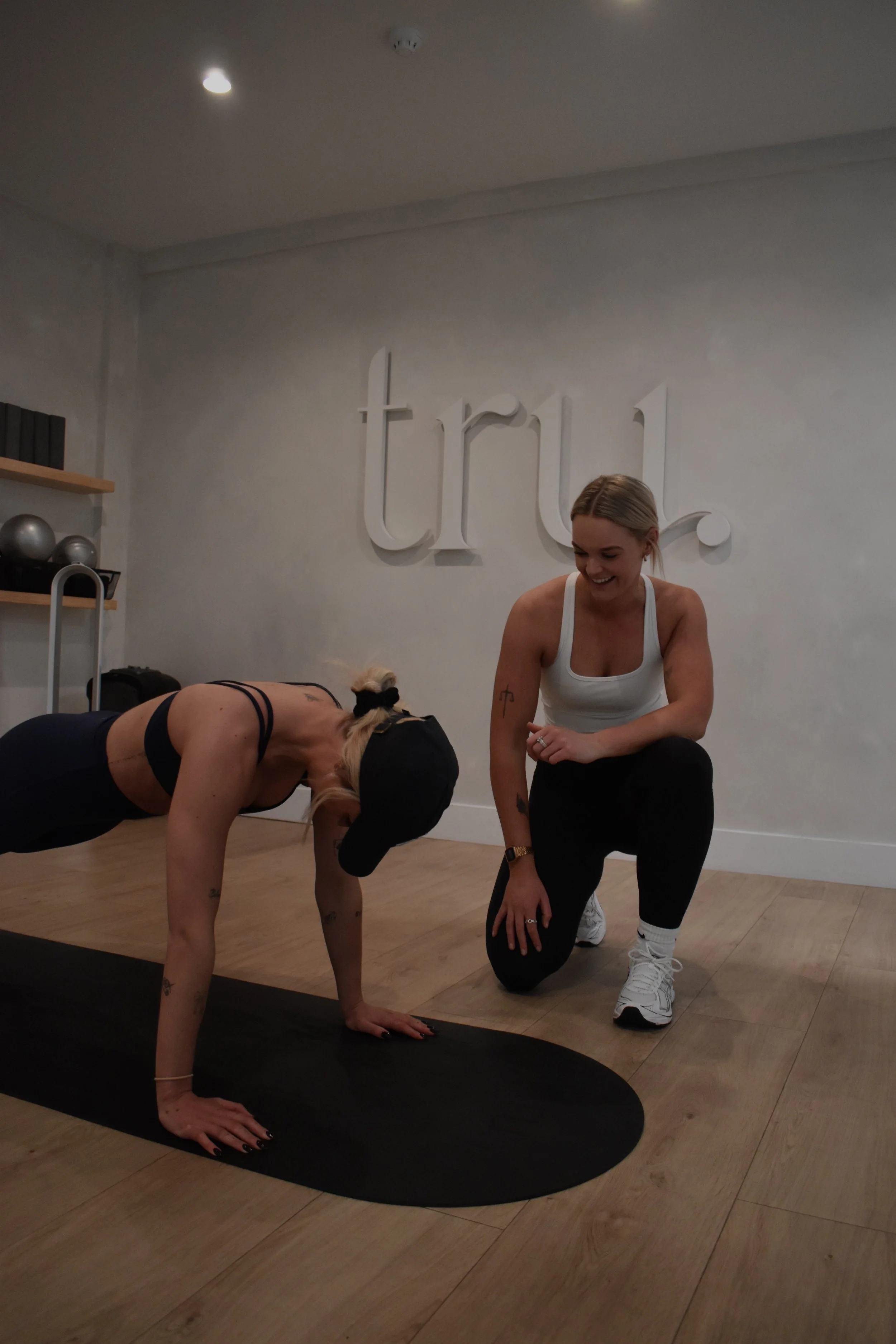 A woman in workout attire is doing a plank on a yoga mat, while a fitness instructor kneels beside her, smiling and offering assistance during a fitness class in a studio with the word 'truth' on the wall.