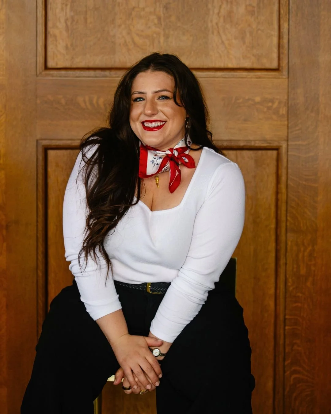 A woman with long dark hair, wearing a white top, black pants, a red and white neck scarf, and red lipstick, smiling and sitting in front of a wood-paneled wall.
