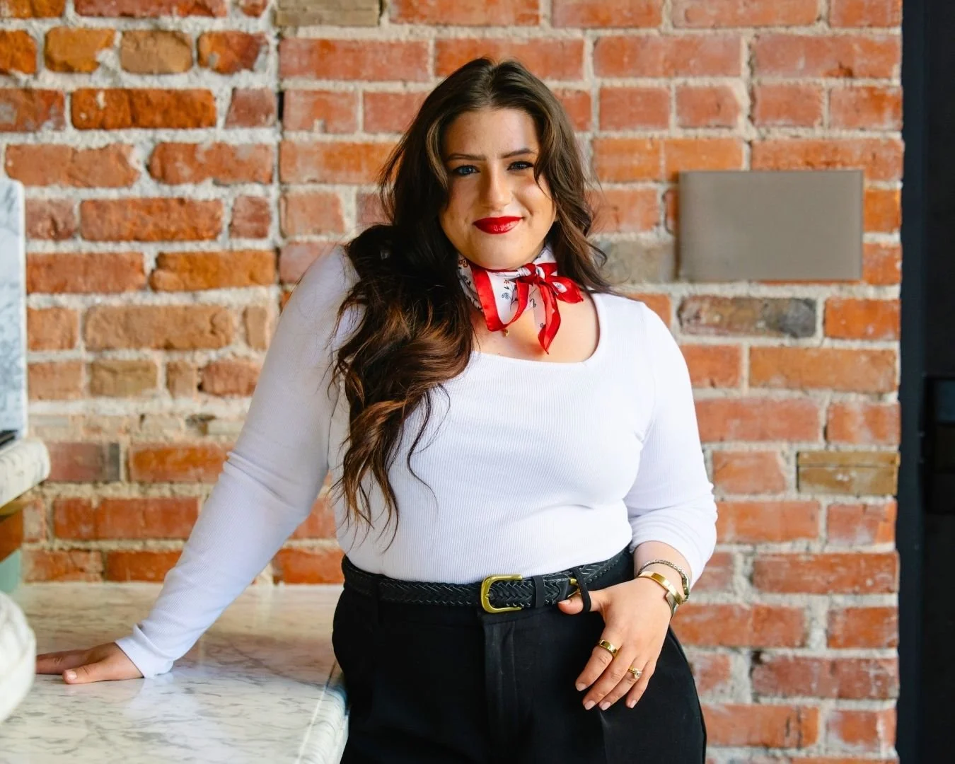 A woman with long brown hair and red lipstick is standing in front of a brick wall. She is wearing a white long sleeve top, a red patterned bandana around her neck, black pants, and a black belt with a yellow buckle. She is leaning on a marble countertop with her left hand resting on her hip and her right hand on the counter, smiling at the camera.