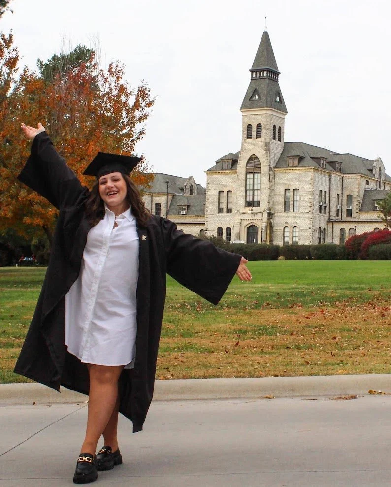 A young woman in a graduation gown and cap standing outside with arms outstretched, in front of a large historic building with a tower, trees with fall foliage, and a grassy lawn.