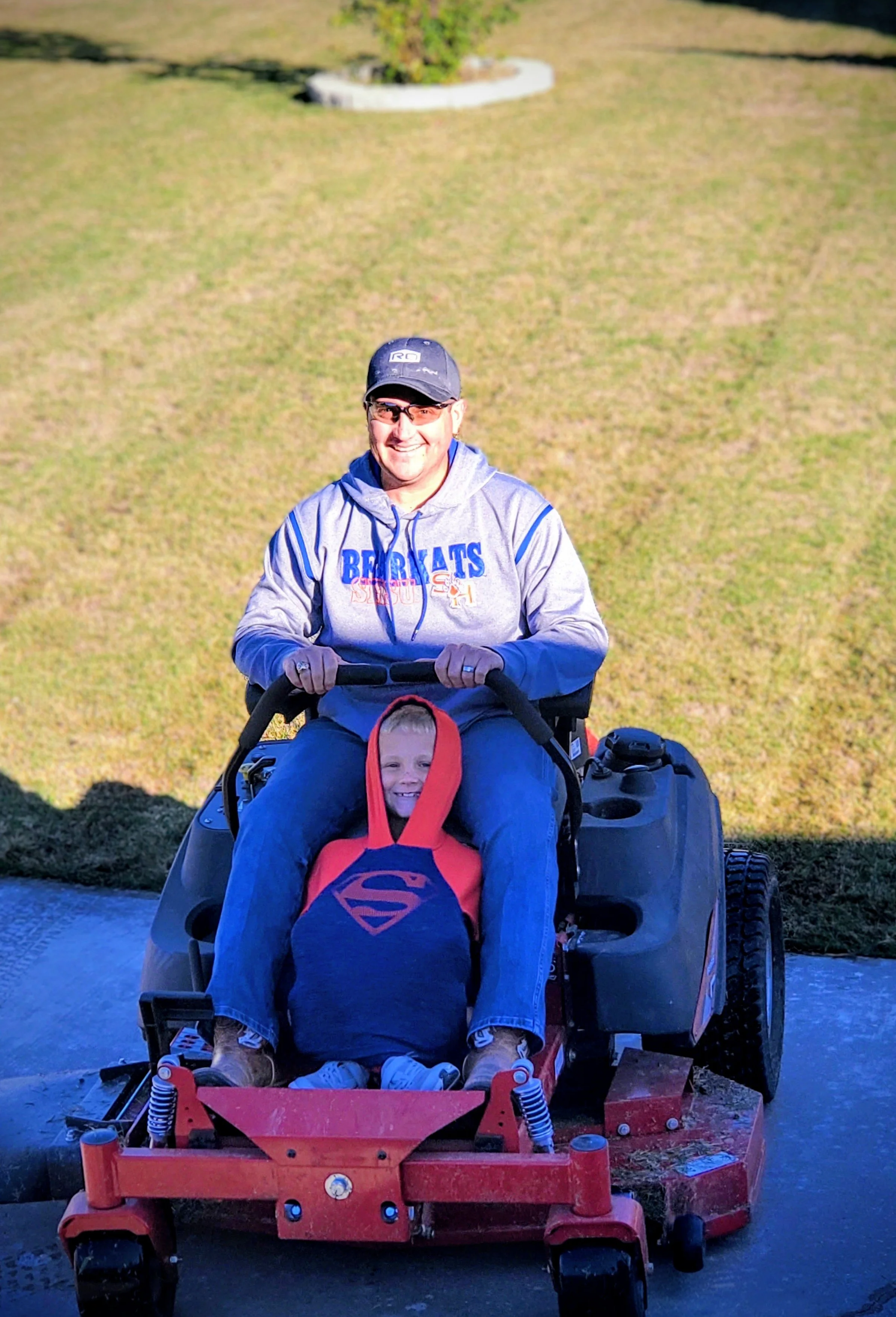 A man and a young boy sitting on a red riding lawn mower outdoors. The man is smiling, wearing a baseball cap, sunglasses, and a gray hoodie with blue lettering. The boy is dressed as Superman, with a noticeable Superman logo on his chest, and is also smiling.