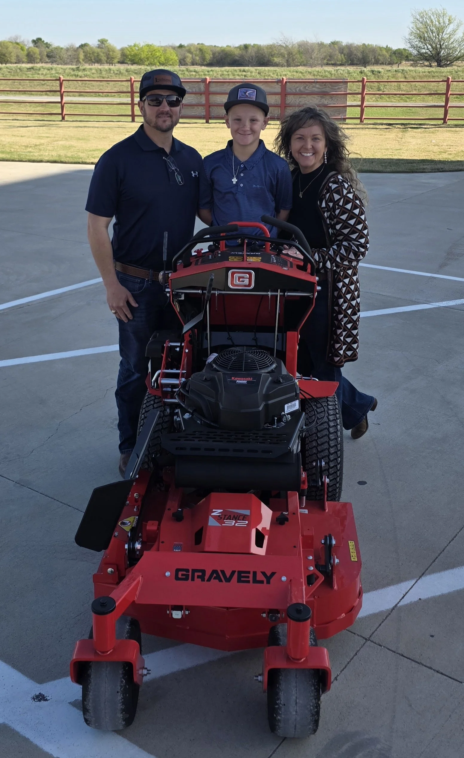 Three people, two men and one woman, standing behind a large red mower with a young boy in the middle. All are smiling outdoors on a paved surface, with a grassy field and trees in the background.