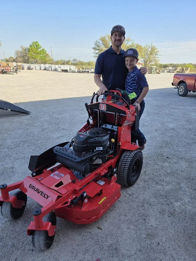 A man and a boy are standing together outdoors, with the boy sitting on a red zero-turn mower. The man has his arm around the boy, and both are smiling. The background shows an open area with some trees, a few vehicles, and a clear blue sky.