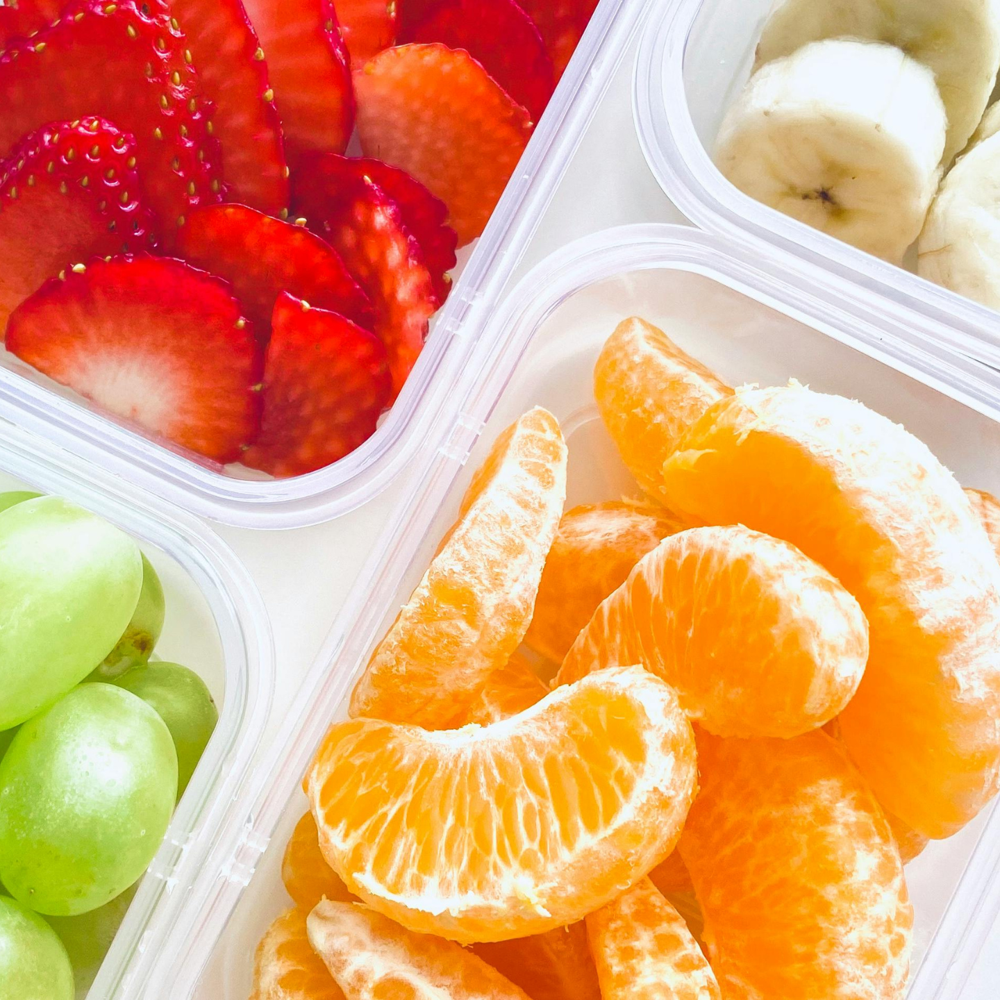 Assorted fresh fruit in clear containers, including sliced strawberries, banana slices, green grapes, and peeled orange segments.