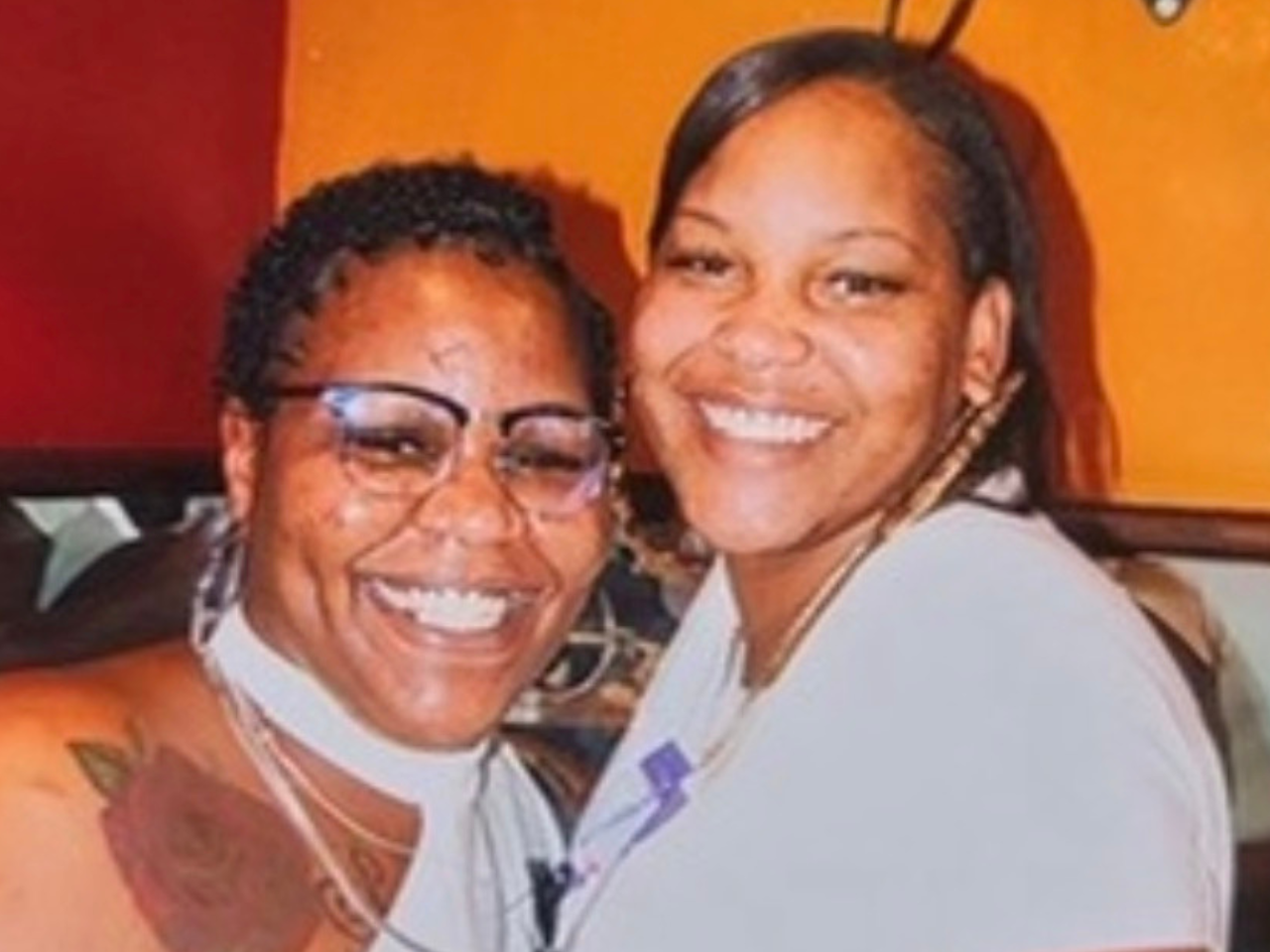Two women smiling and embracing each other indoors with an orange wall in the background.