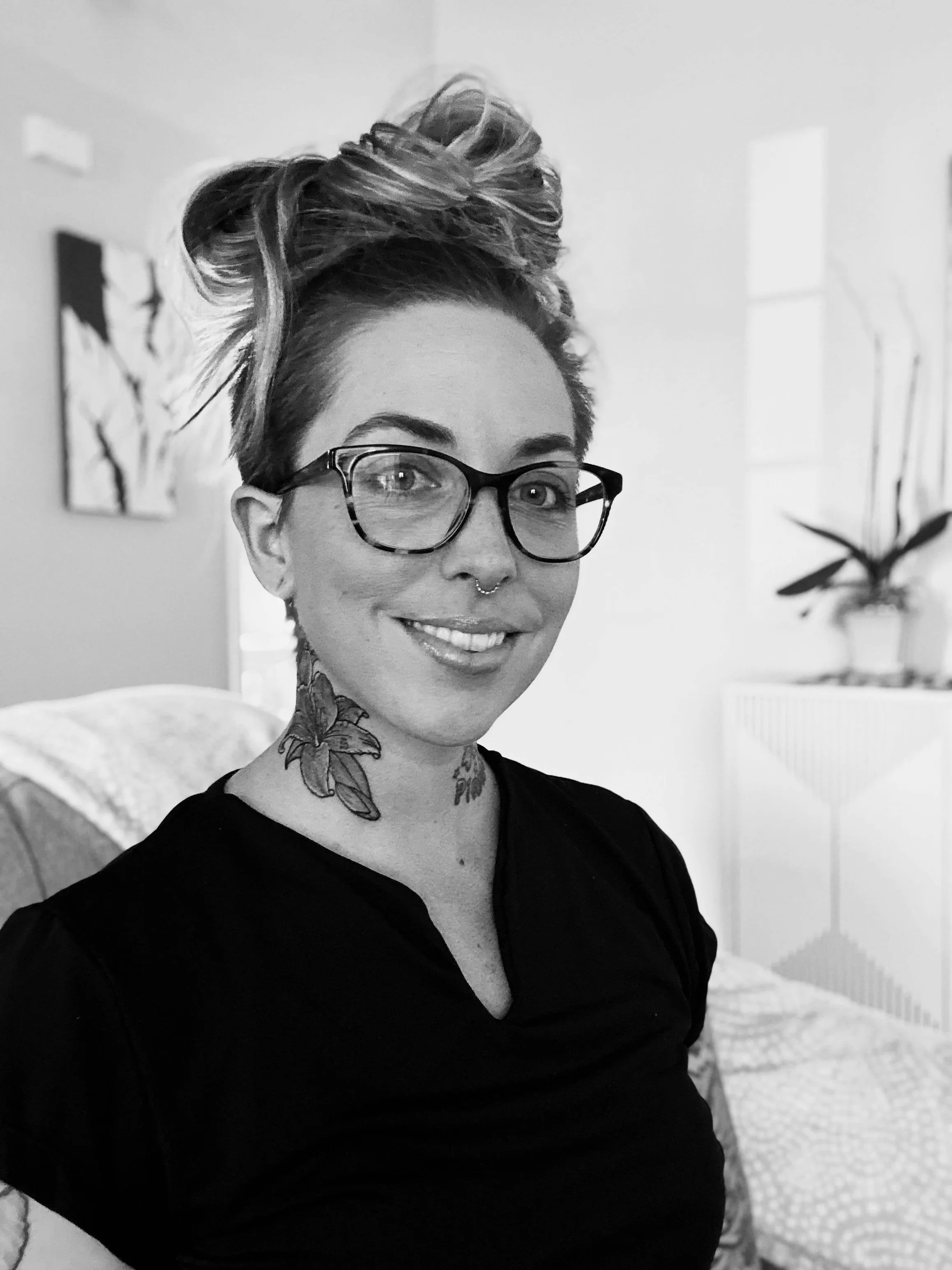 Black and white photo of a woman with glasses, tattoos, and a messy bun smiling in a cozy room with a couch, plants, and artwork.