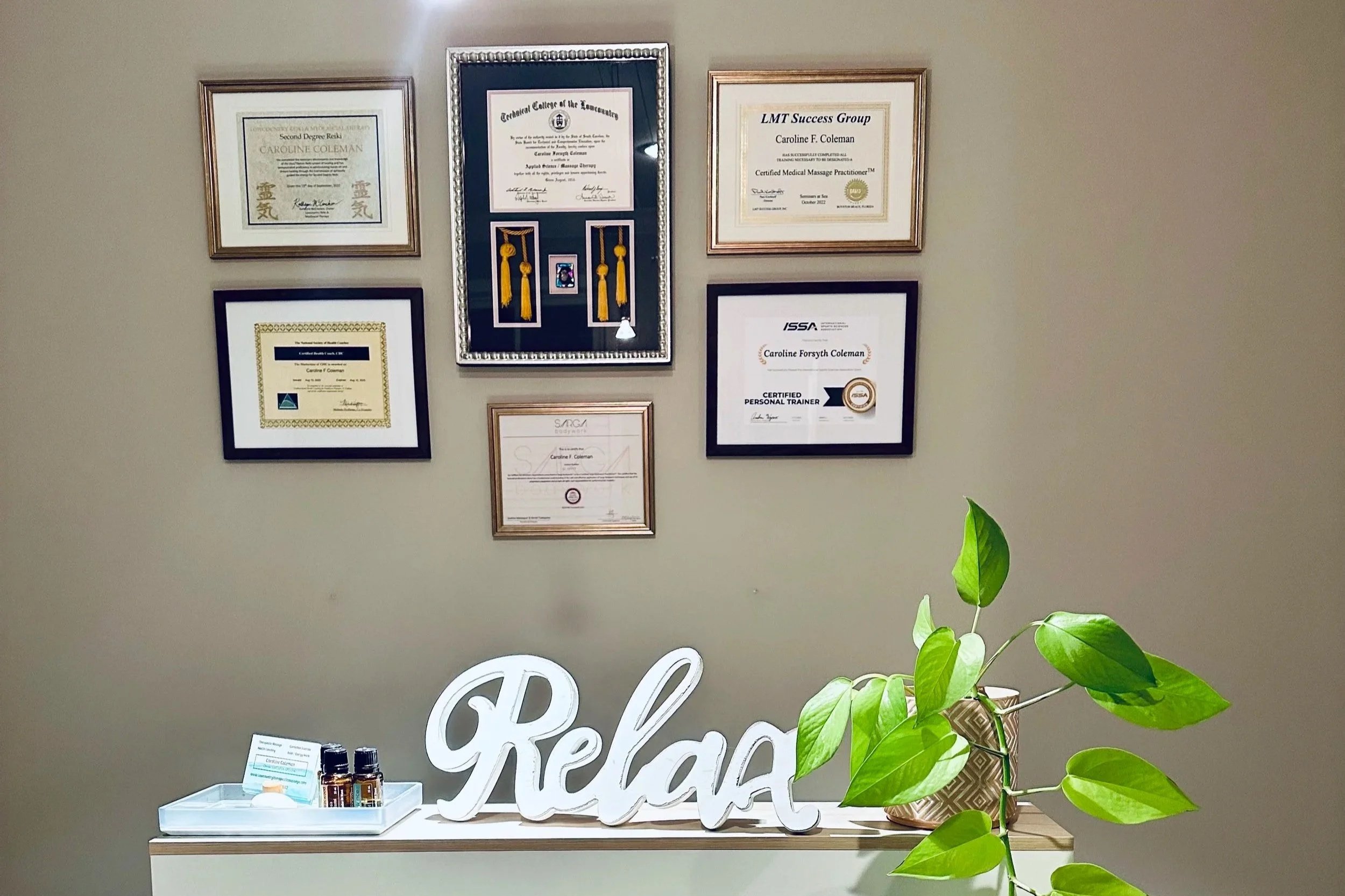 Display of framed certificates and diplomas on a beige wall, a white decorative 'relax' sign, a table with small bottles, and a green leafy plant in the foreground.