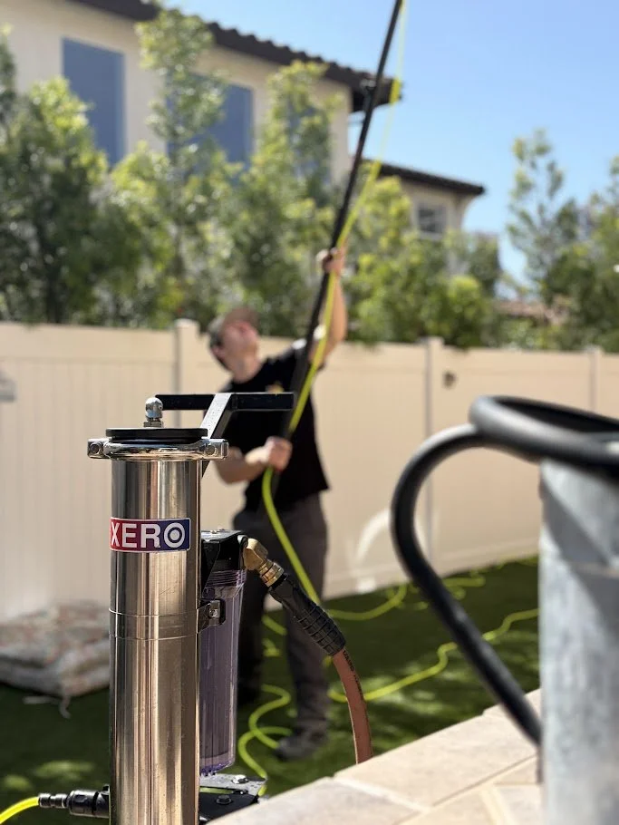 A person holding a long yellow pole with a black attachment, possibly applying or cleaning with water or other liquid, in a backyard with a white fence, house, green trees, and a pillow on the grass.