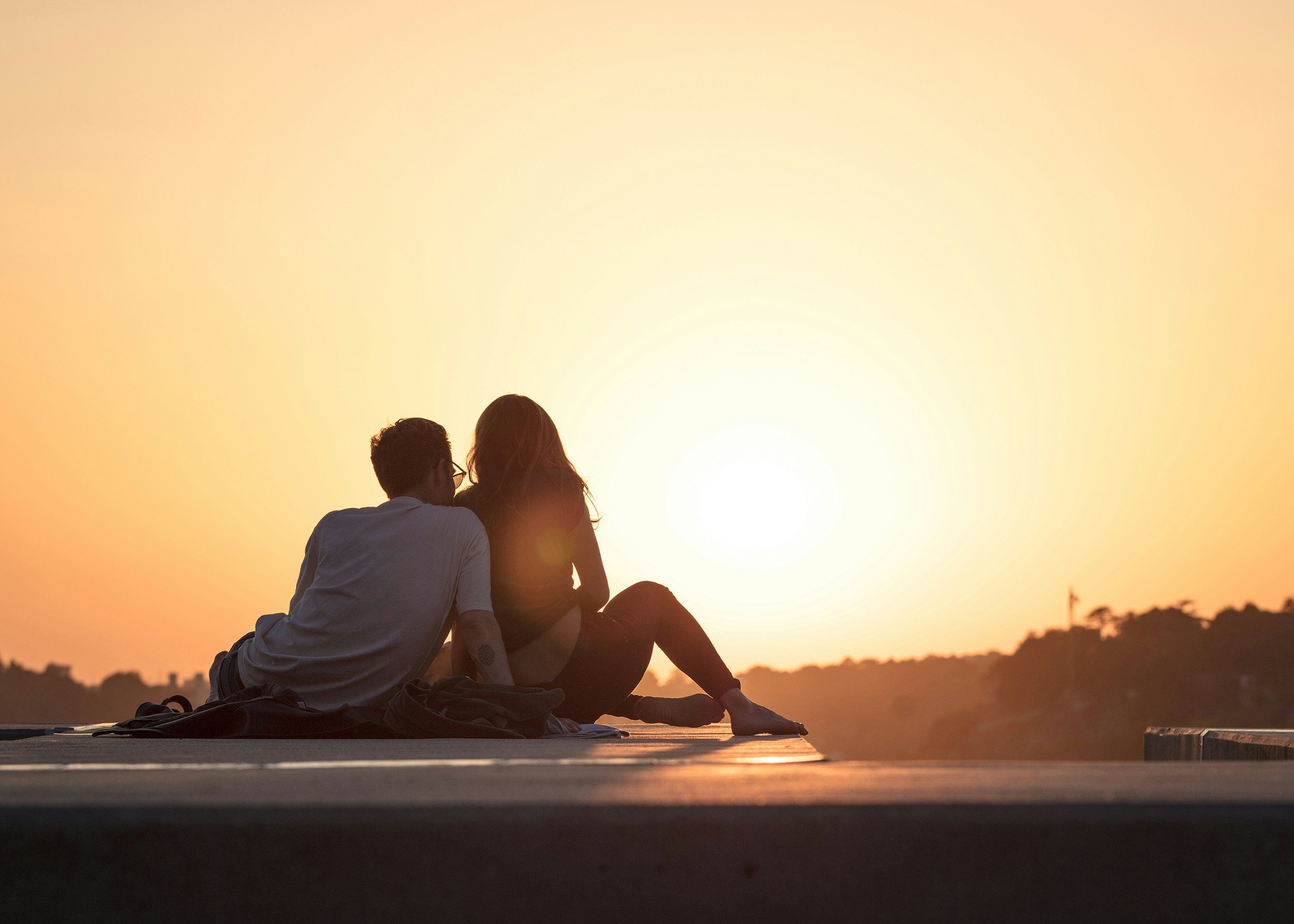 A couple sitting together outdoors at sunset, with a warm yellow-orange sky and a scenic landscape in the background.
