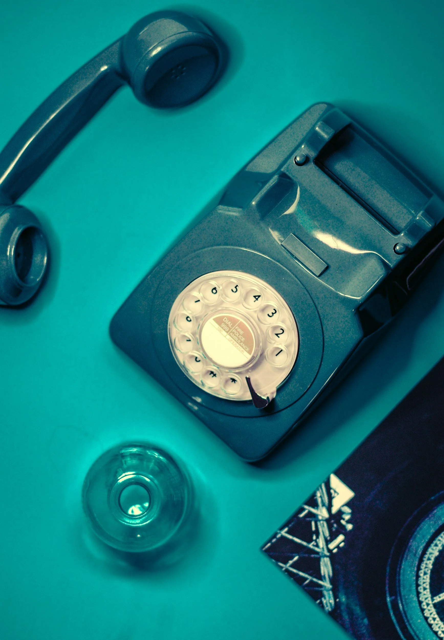 A vintage gray rotary telephone with a white dial and black numbers on a teal surface, alongside a clear glass and a printed photograph.