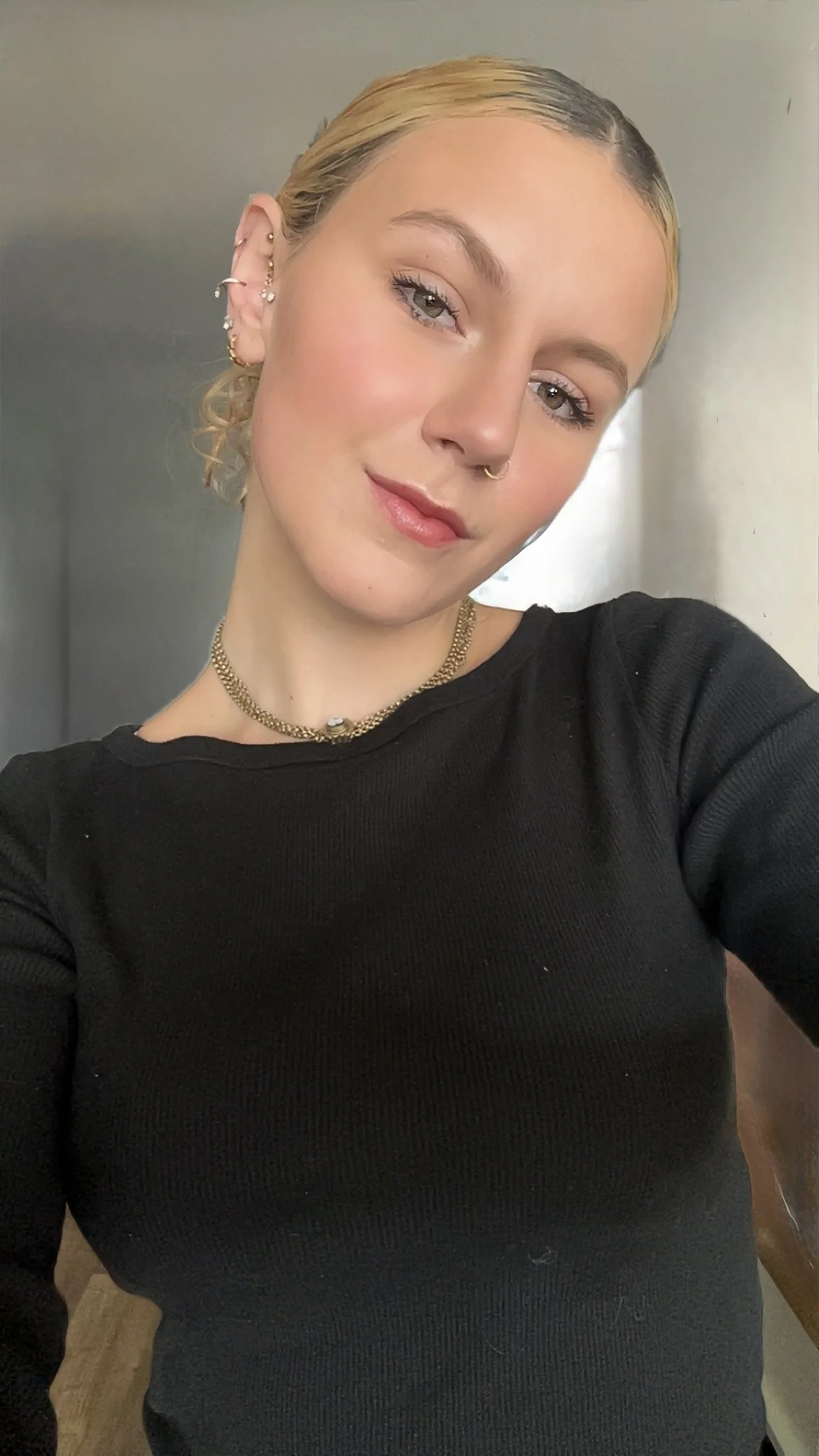 A young woman with blonde hair in a slicked-back style, wearing gold jewelry including earrings, a necklace, and a septum ring, takes a selfie indoors with a neutral background, wearing a black long-sleeve top.