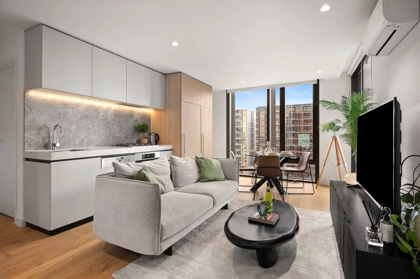 Modern living room and kitchen with beige sofa, black coffee table, television, dining table with four chairs, large windows showing city buildings, potted plants, and wooden flooring.