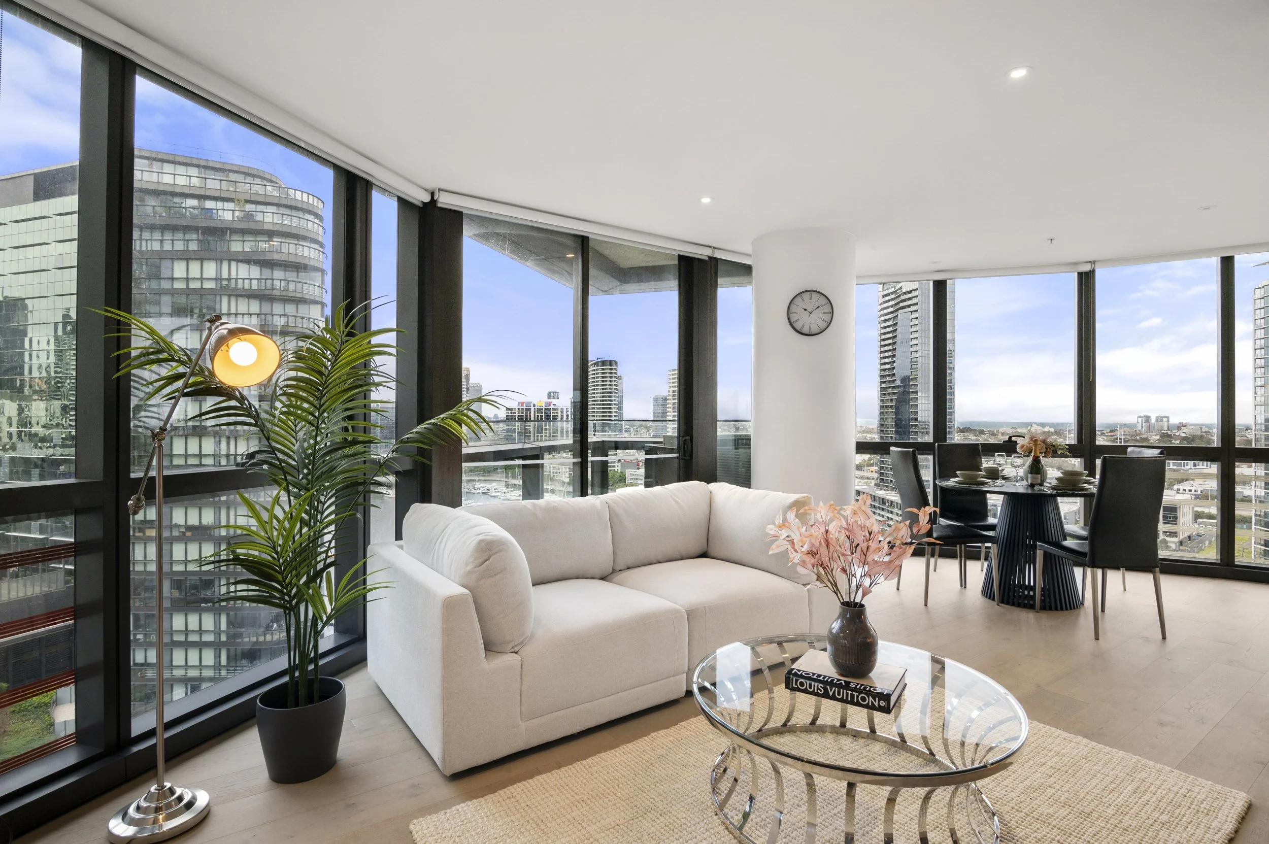 Modern living room with large floor-to-ceiling windows, white sofa, glass coffee table with pink flowers, potted plant, dining area with black chairs, and city skyline view