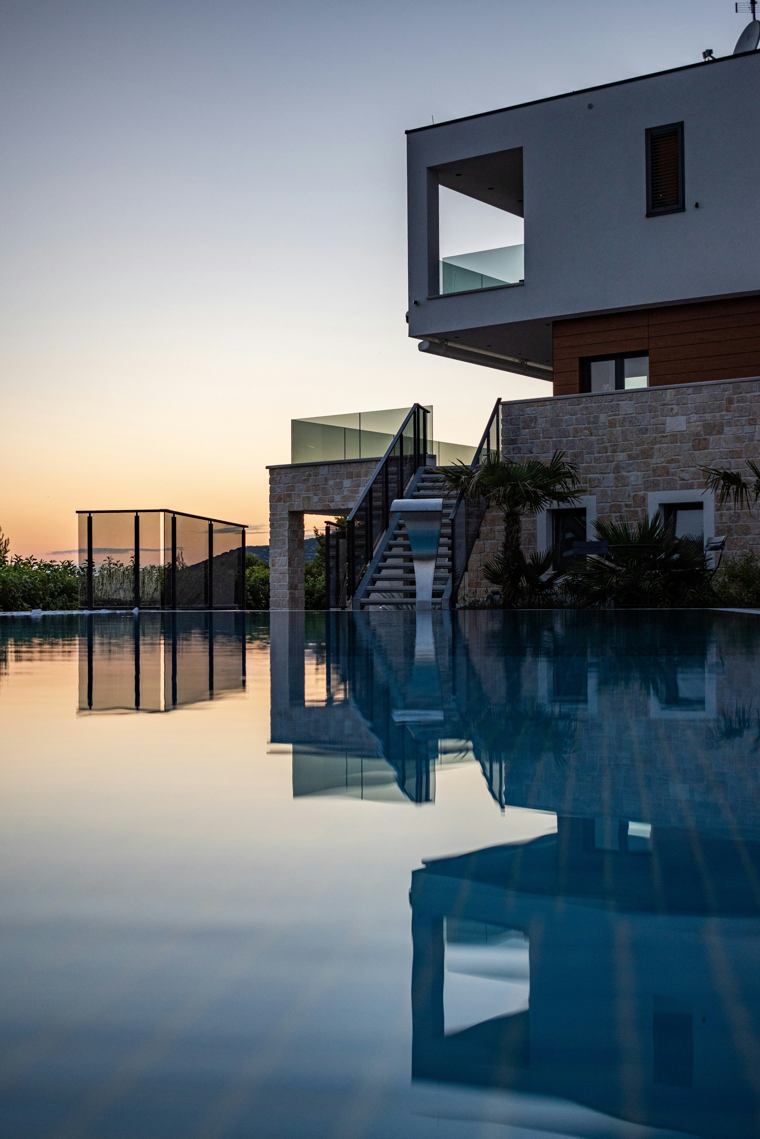 Modern house with a staircase and a glass balcony overlooking a swimming pool at sunset.