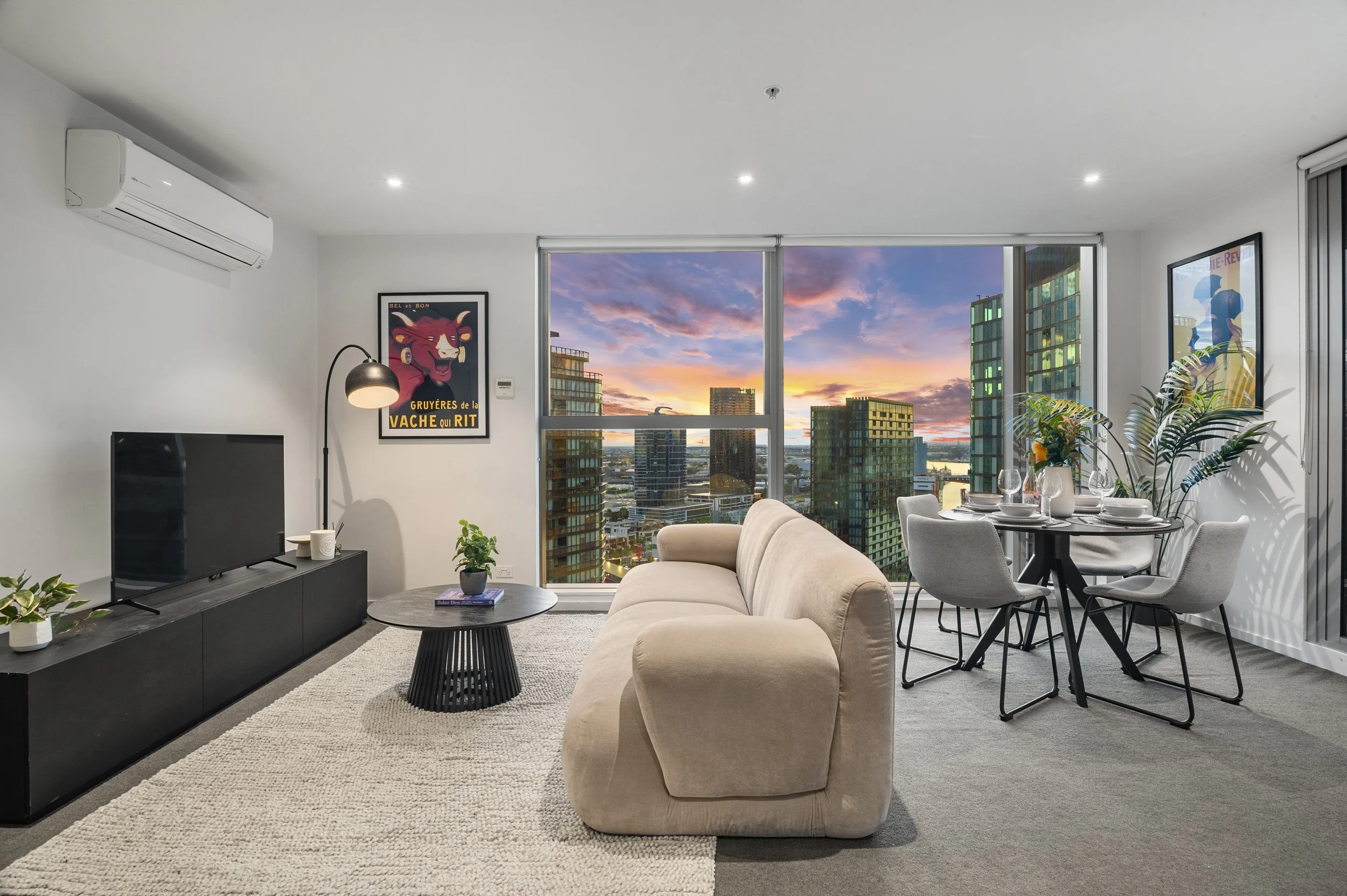Living room with beige sofa, black TV stand, flat screen TV, round black coffee table, gray dining table with four chairs, large windows with city skyline at sunset, wall art, plants, and a textured rug.
