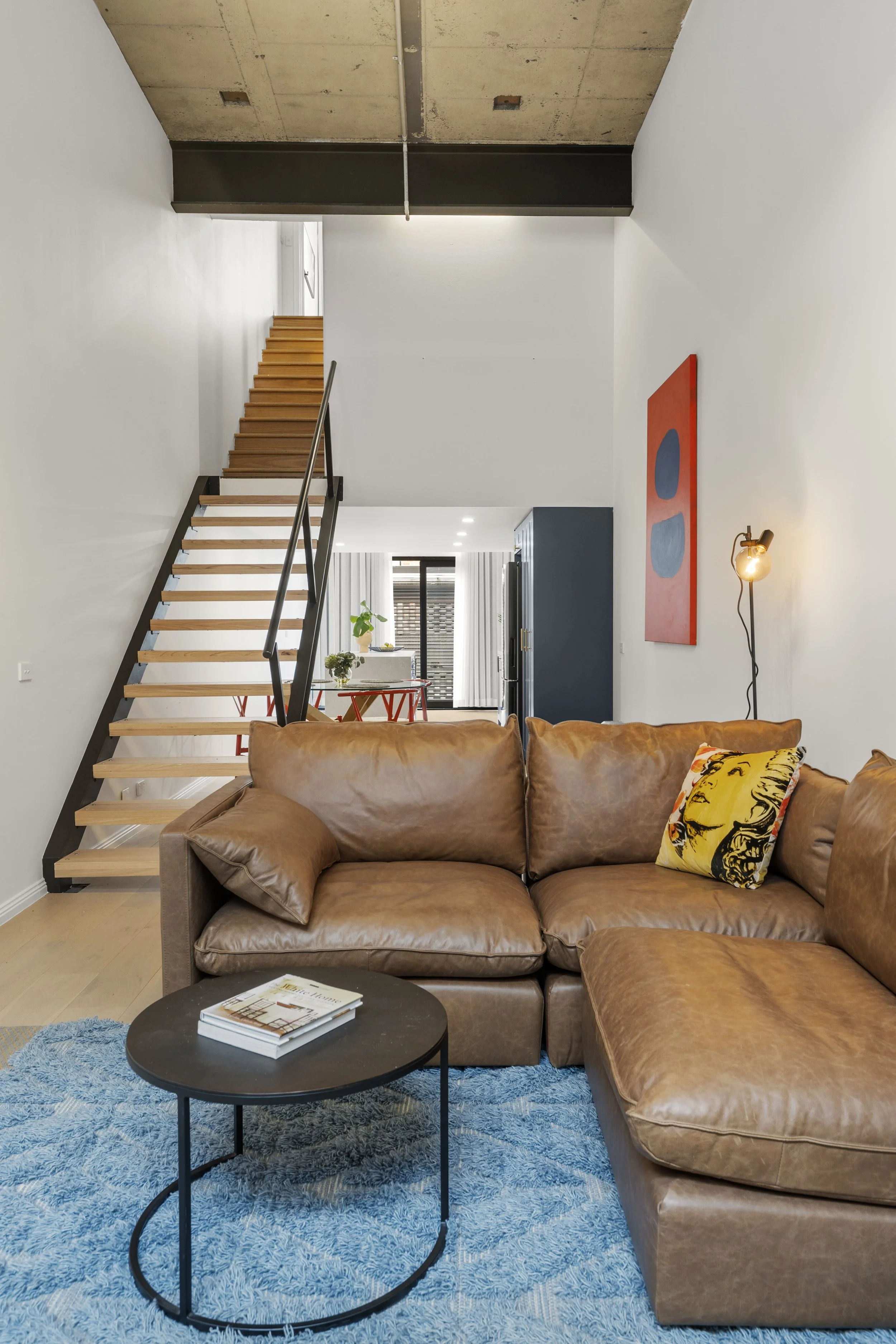 Living room with a brown leather sectional sofa on a blue rug, a black round coffee table with magazines, a modern wall lamp, a colorful abstract wall art, a staircase with wooden steps and black railing, visible in an open-plan design that leads to a dining and kitchen area with natural light.