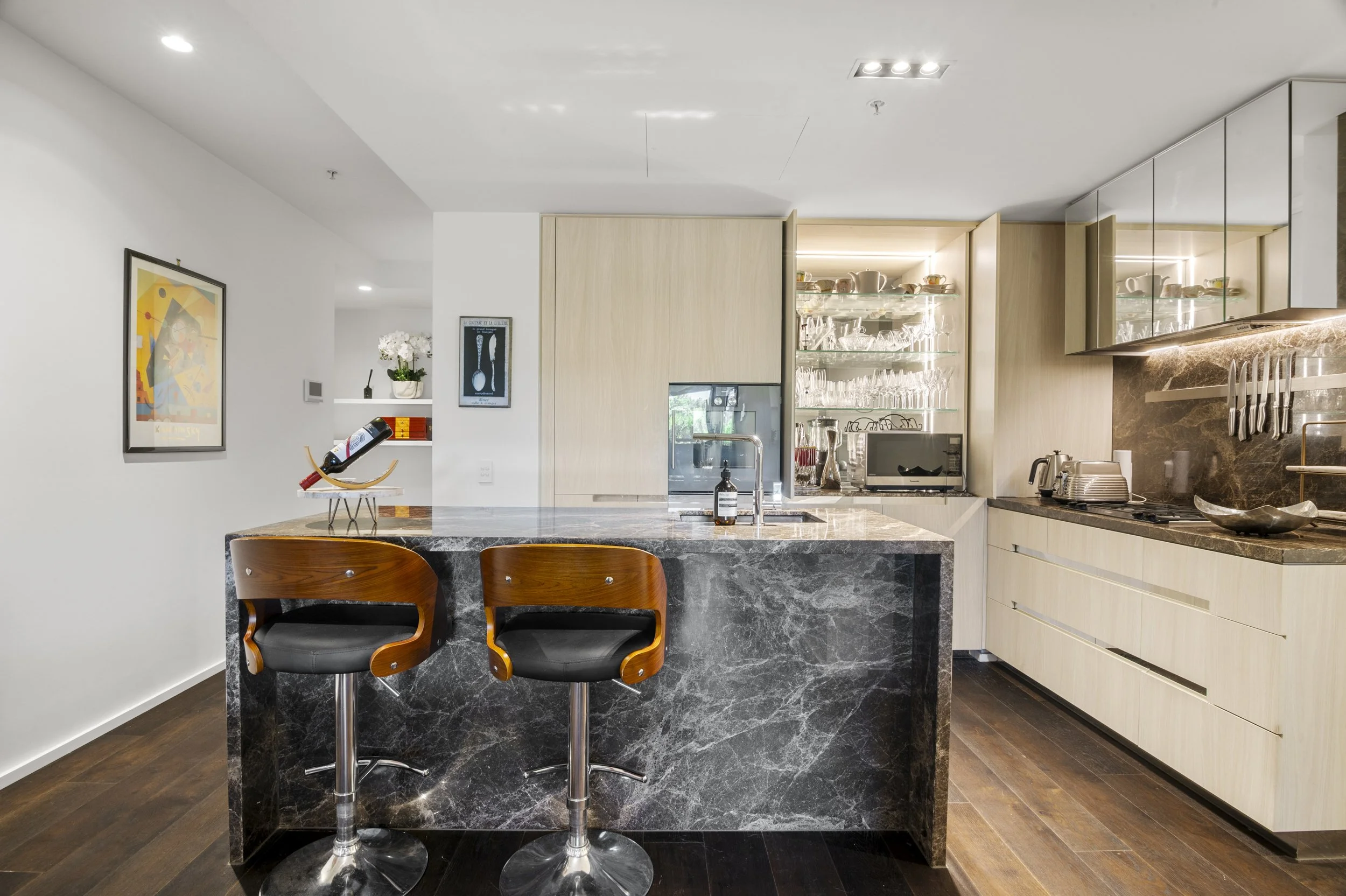Modern kitchen with a marble island, two wooden bar stools, glassware display, and kitchen appliances.
