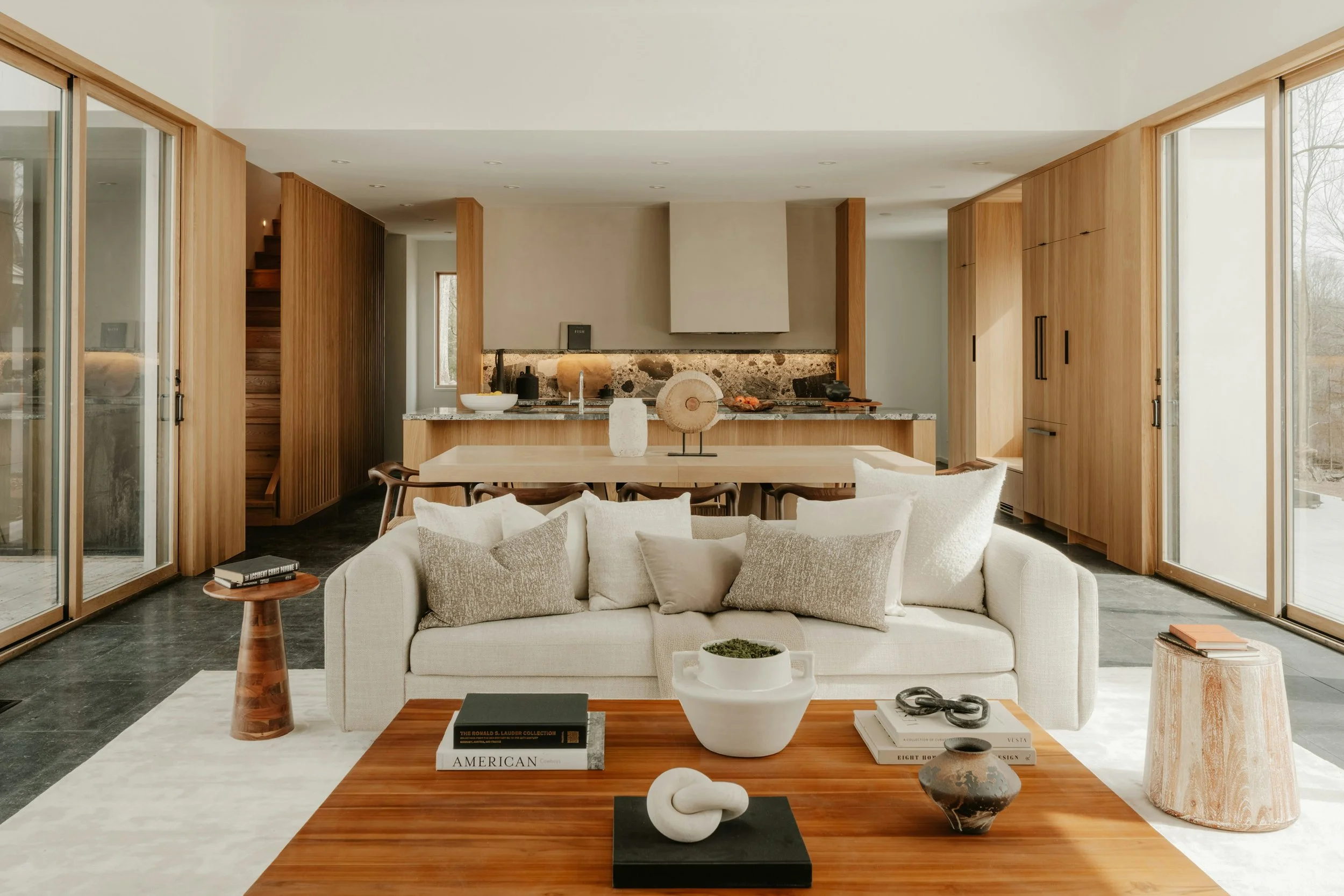 Living room with cream sofa, wooden coffee table, decorative objects, and large glass sliding doors leading outside. Open kitchen with wooden cabinetry and stone backsplash in the background.