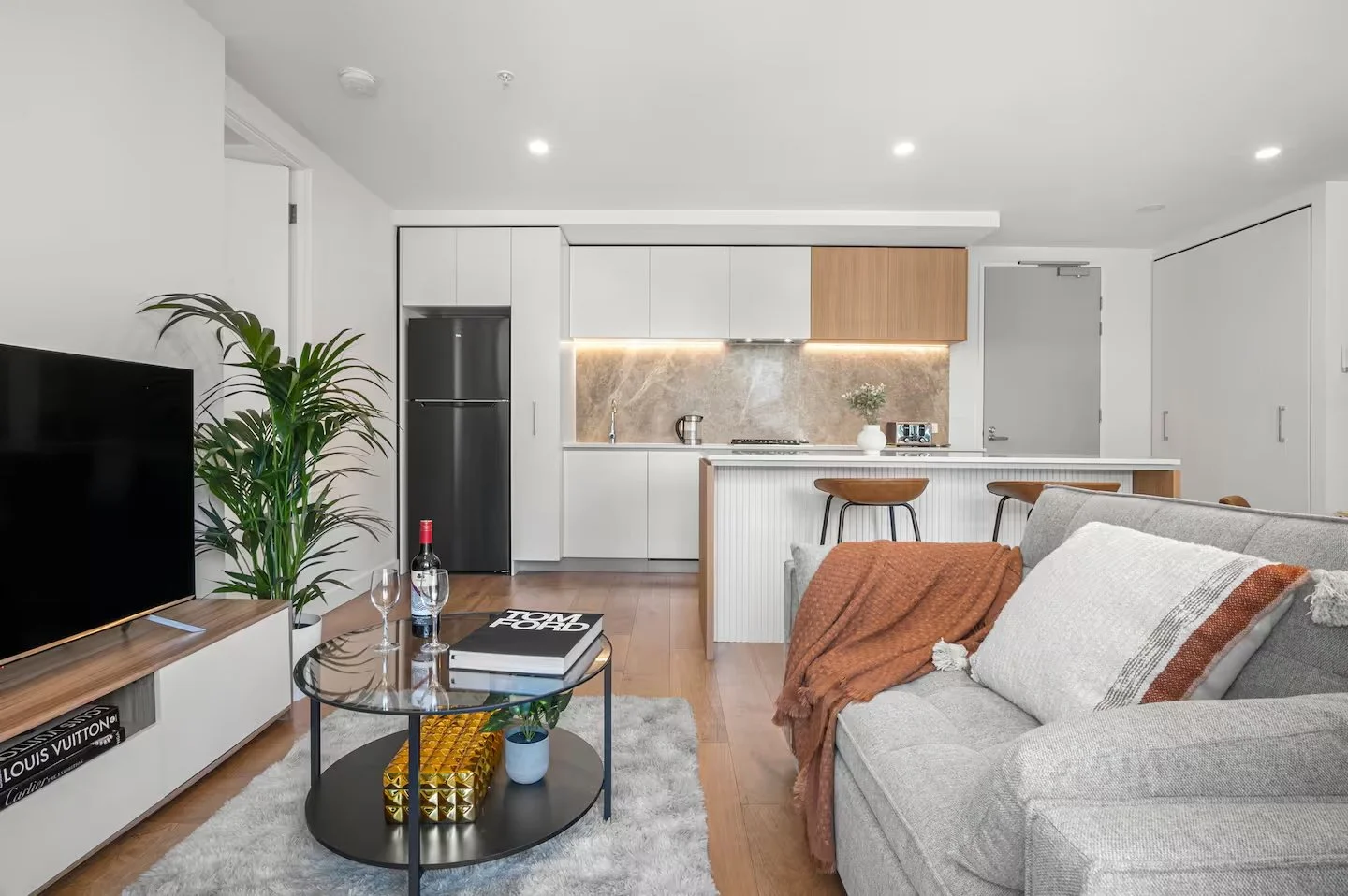 Modern open-concept living room and kitchen with white cabinets, gray refrigerator, wooden accents, cozy sofa with orange throw, coffee table with wine and books, flat-screen TV, and decorative plants.