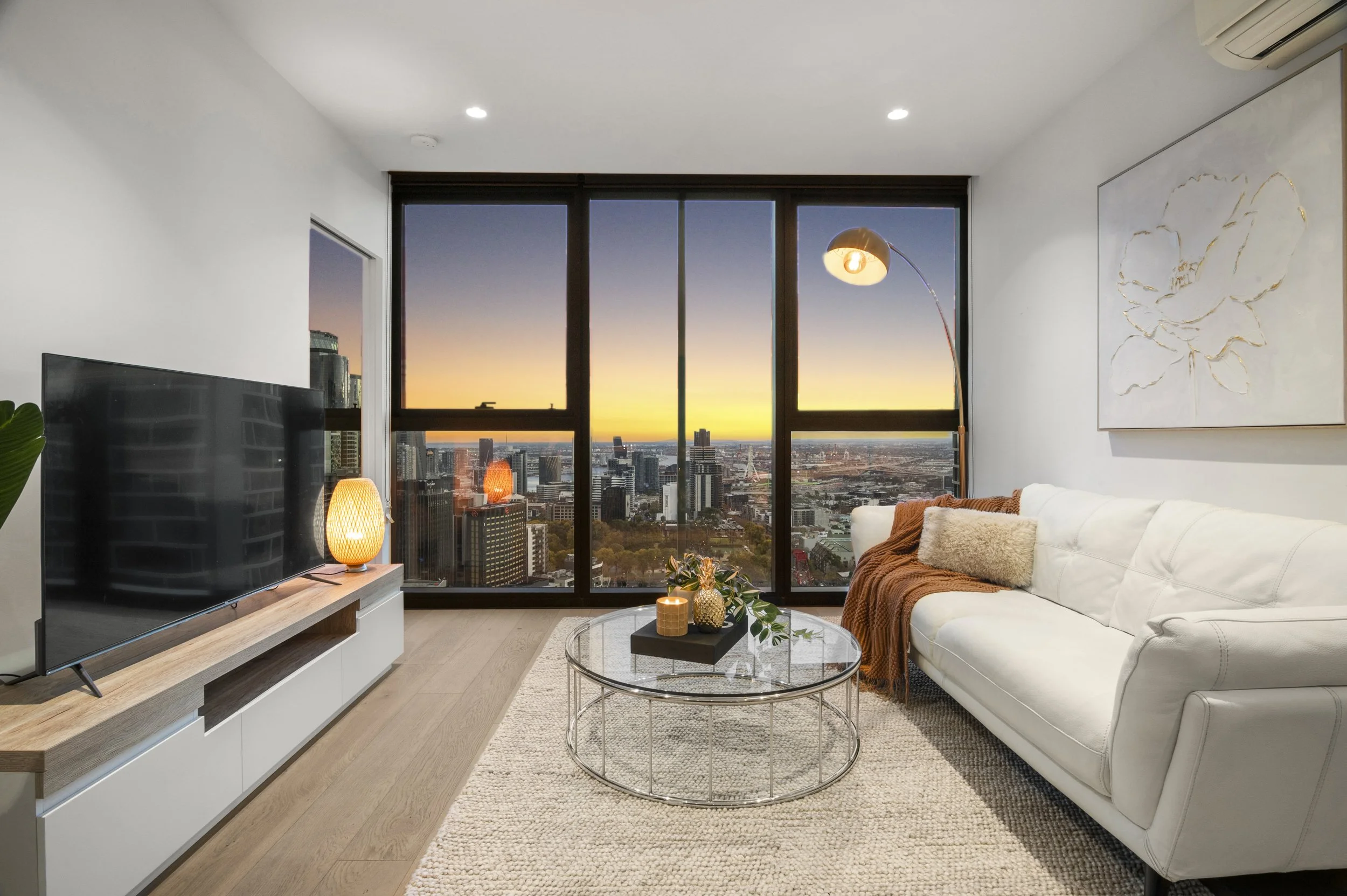 Modern living room with large floor-to-ceiling windows showing a cityscape and sunset. Features a white sofa with brown blanket and beige pillow, a glass coffee table with decorative items, a TV on a wooden stand with a lamp, and a tall floor lamp. White walls with a large floral artwork.