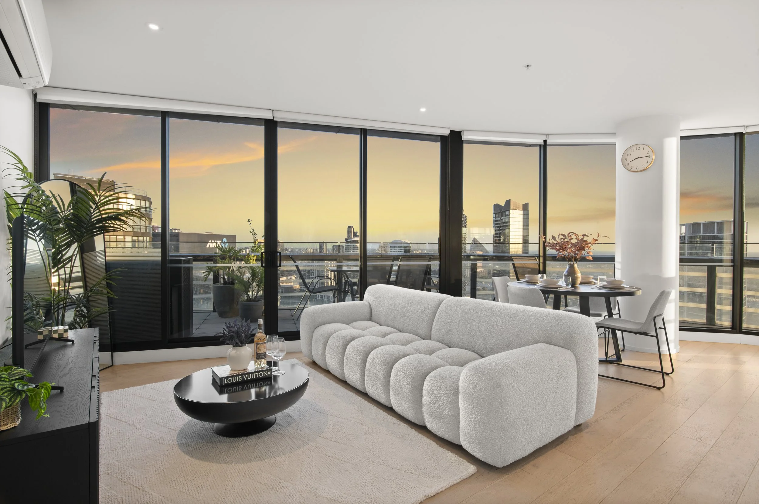 Modern living room with large floor-to-ceiling windows showing a cityscape at sunset, white textured sofa, round black coffee table with books and a bottle, plush beige rug, potted plants, and a dining table with chairs in the background.