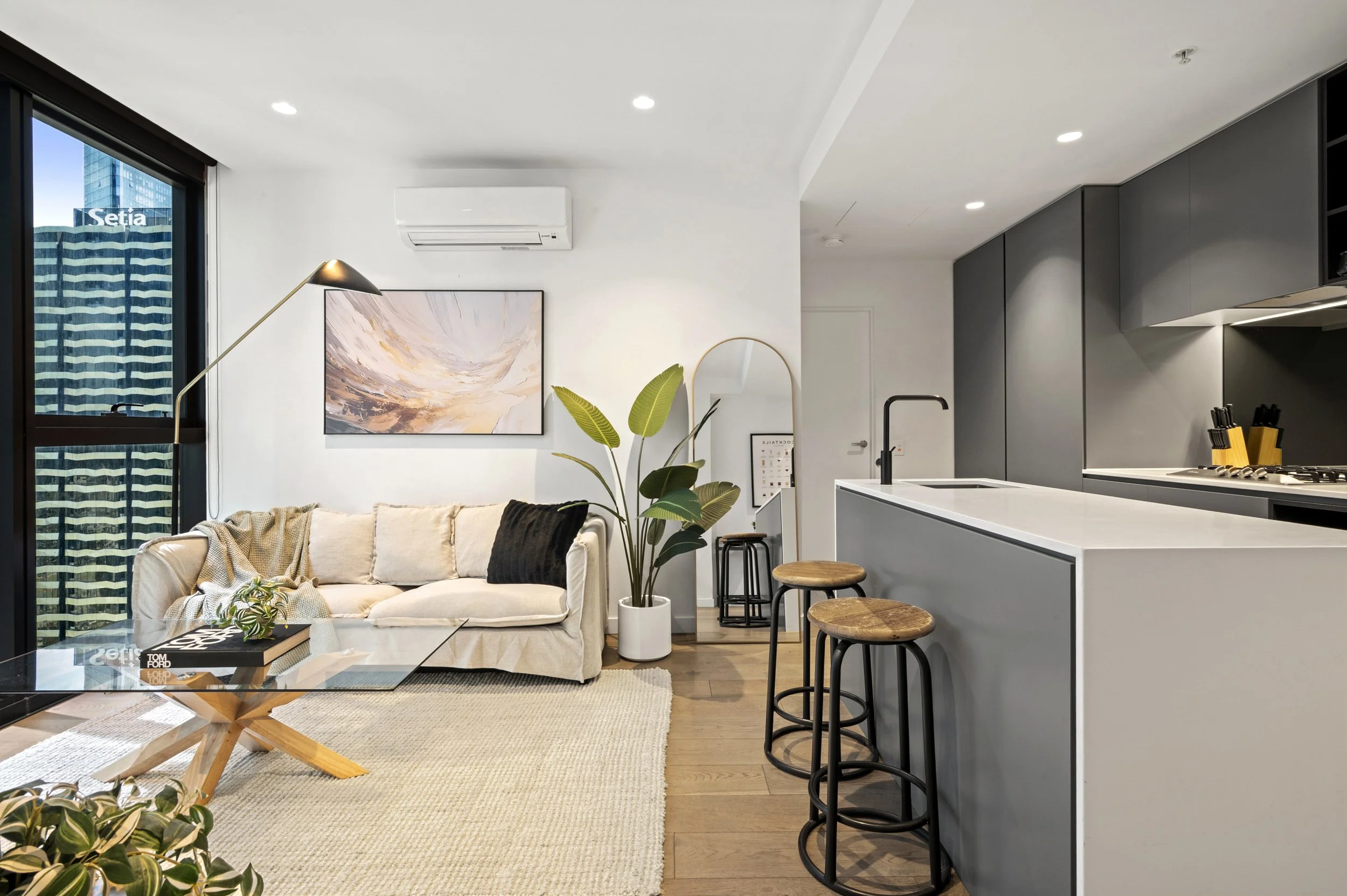 Modern open-concept living room and kitchen with white walls, a beige sofa, black and wooden barstools, a large window, a potted plant, and minimalist decor.