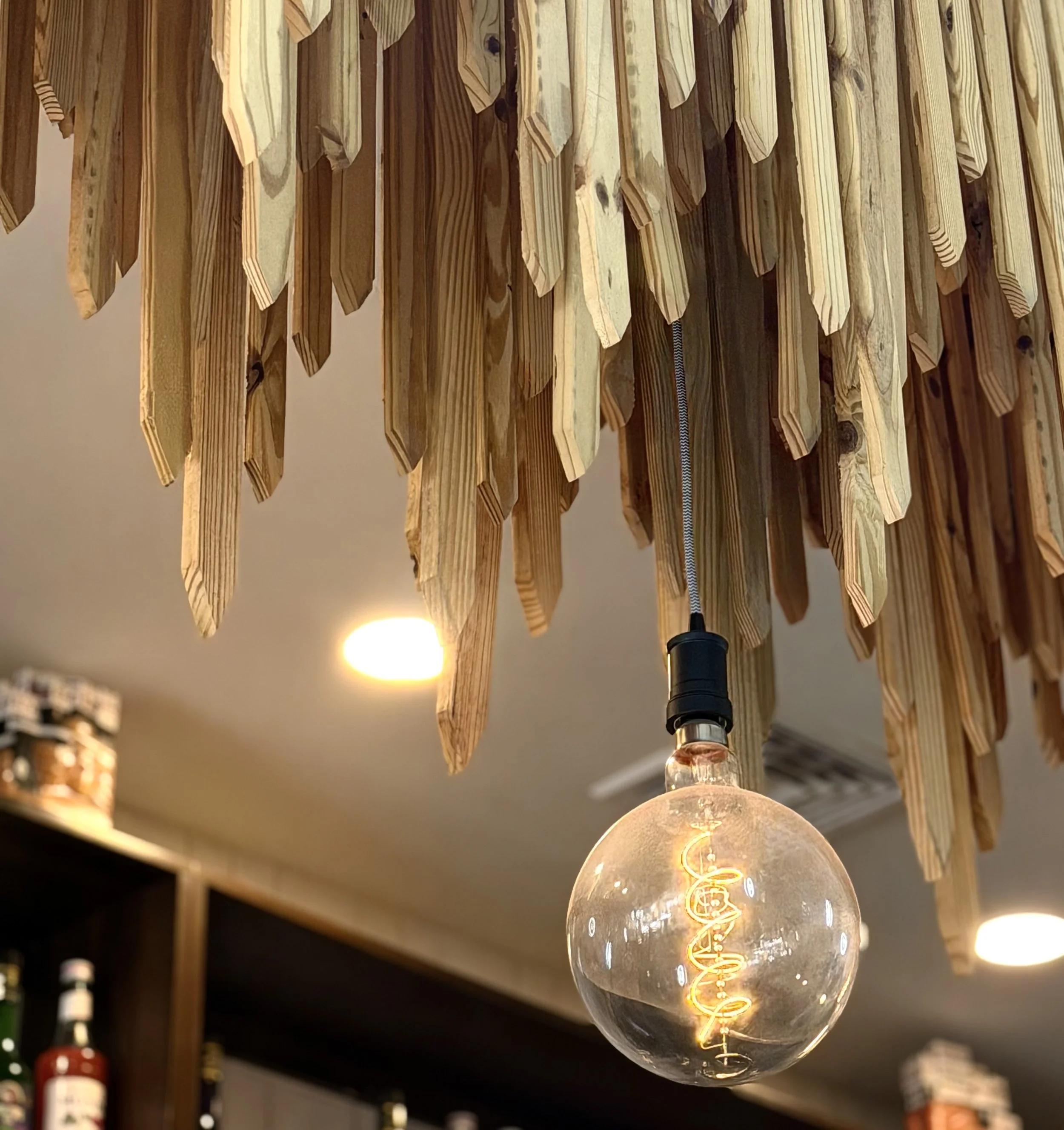 Close-up of an illuminated vintage-style filament light bulb hanging from a ceiling, with a decorative wooden wall with hanging wooden slats in the background.