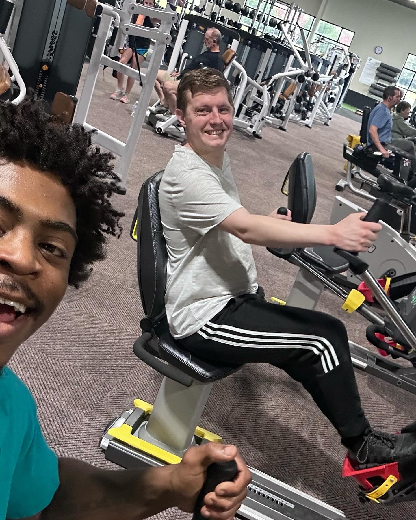 Two people taking a selfie inside a gym. One is smiling in the foreground with curly hair, and the other is sitting on a workout bike, smiling at the camera, wearing a white T-shirt and black track pants with white stripes. The gym background has various exercise equipment.