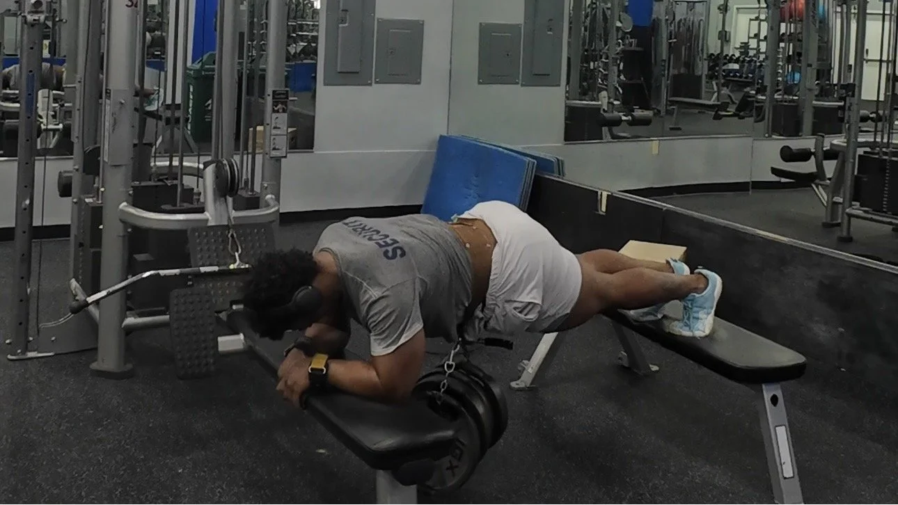 A man performing a plank exercise on an incline bench at a gym, with a weight plate hanging from his ankle and headphones on.