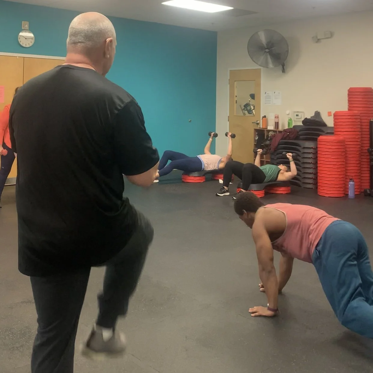A fitness class with multiple women doing exercises with dumbbells and mats, led by an instructor in a gym room with a blue wall, stacked red weights, and a door in the background.